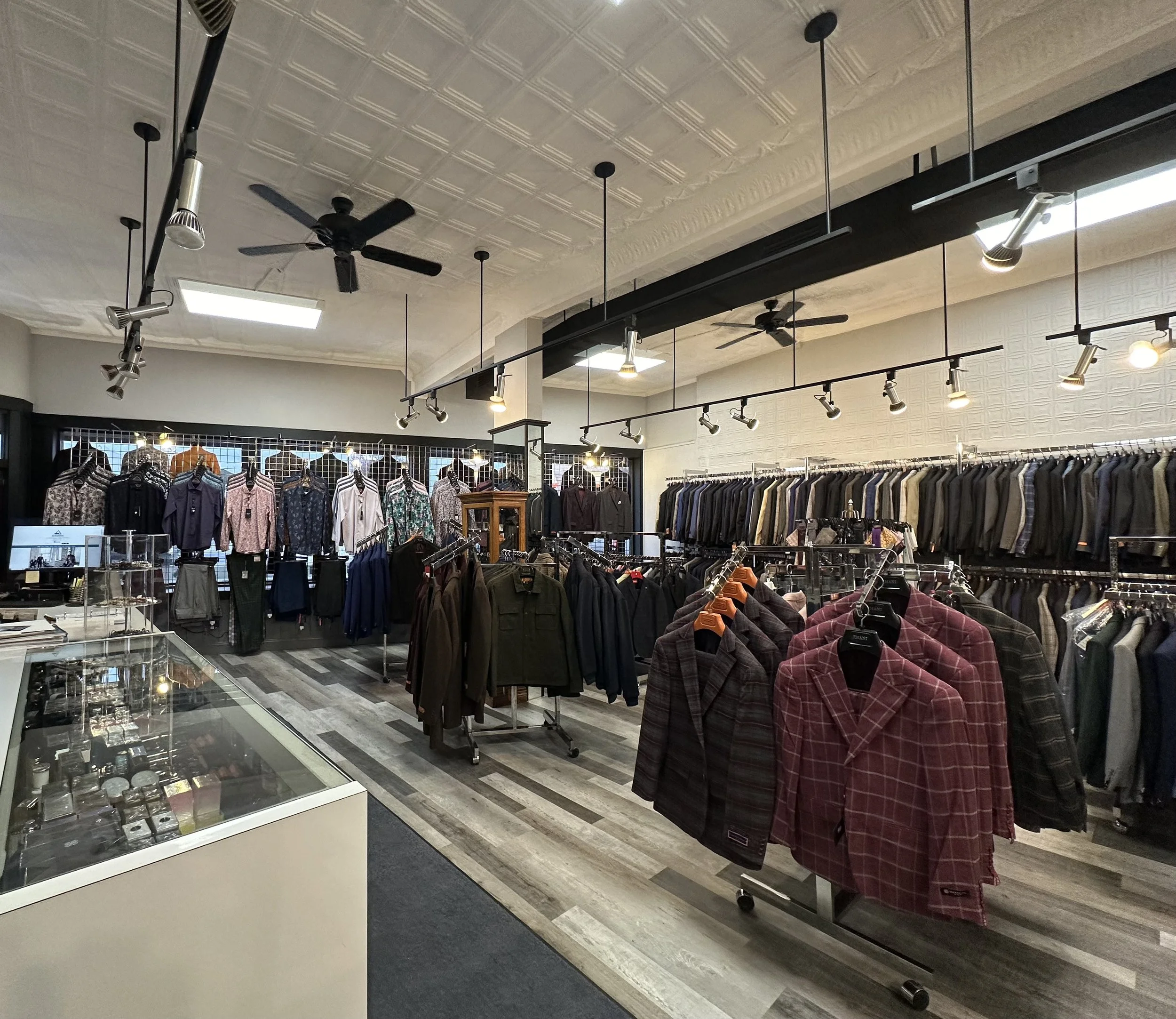 Clothing store interior with racks of shirts, jackets, and suits, white textured walls, track lighting, ceiling fans, and wood-look flooring.