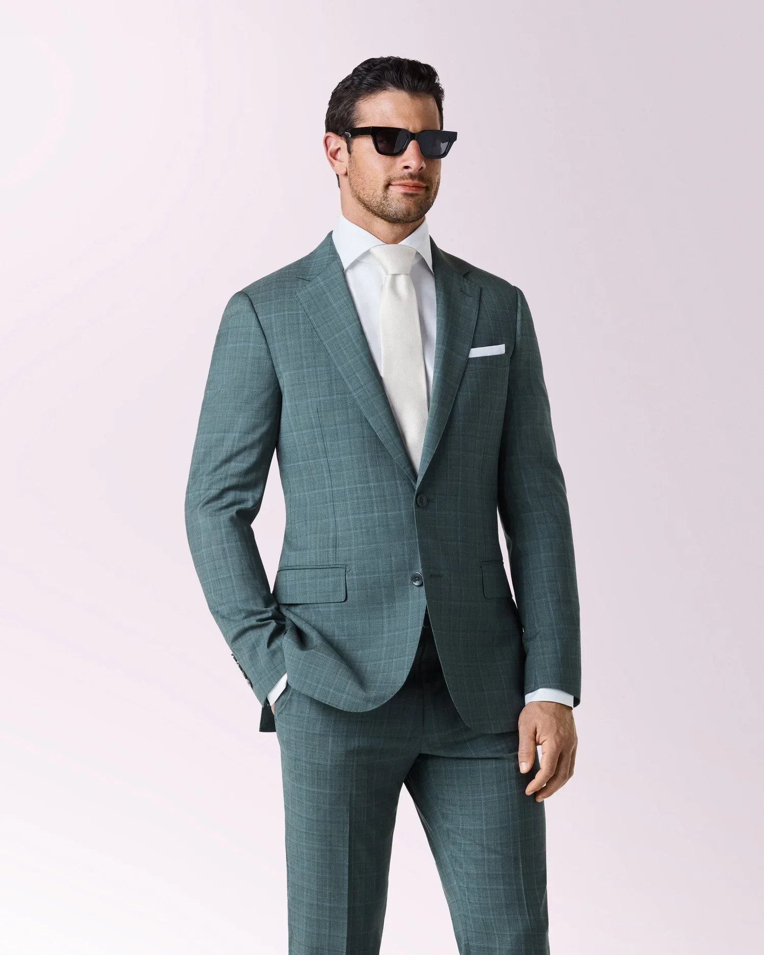 Man in a plaid gray suit, white shirt, white tie, sunglasses, and with one hand in his pocket, standing against a light background.