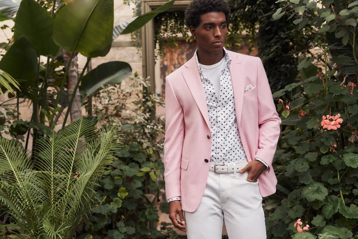 A young man with dark skin and curly hair standing amidst lush green plants. He is wearing a light pink blazer over a white shirt with black polka dots, light-colored pants, and a white belt. He looks contemplative, with one hand in his pocket.