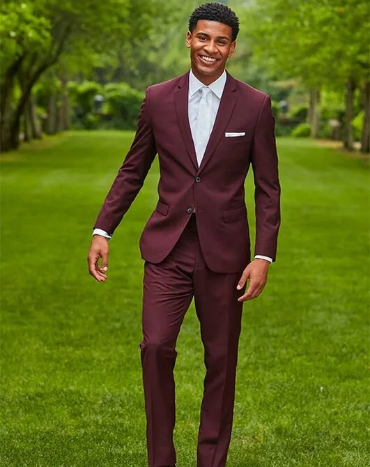 A smiling man in a maroon suit and white shirt walking on a lush green lawn with trees in the background.