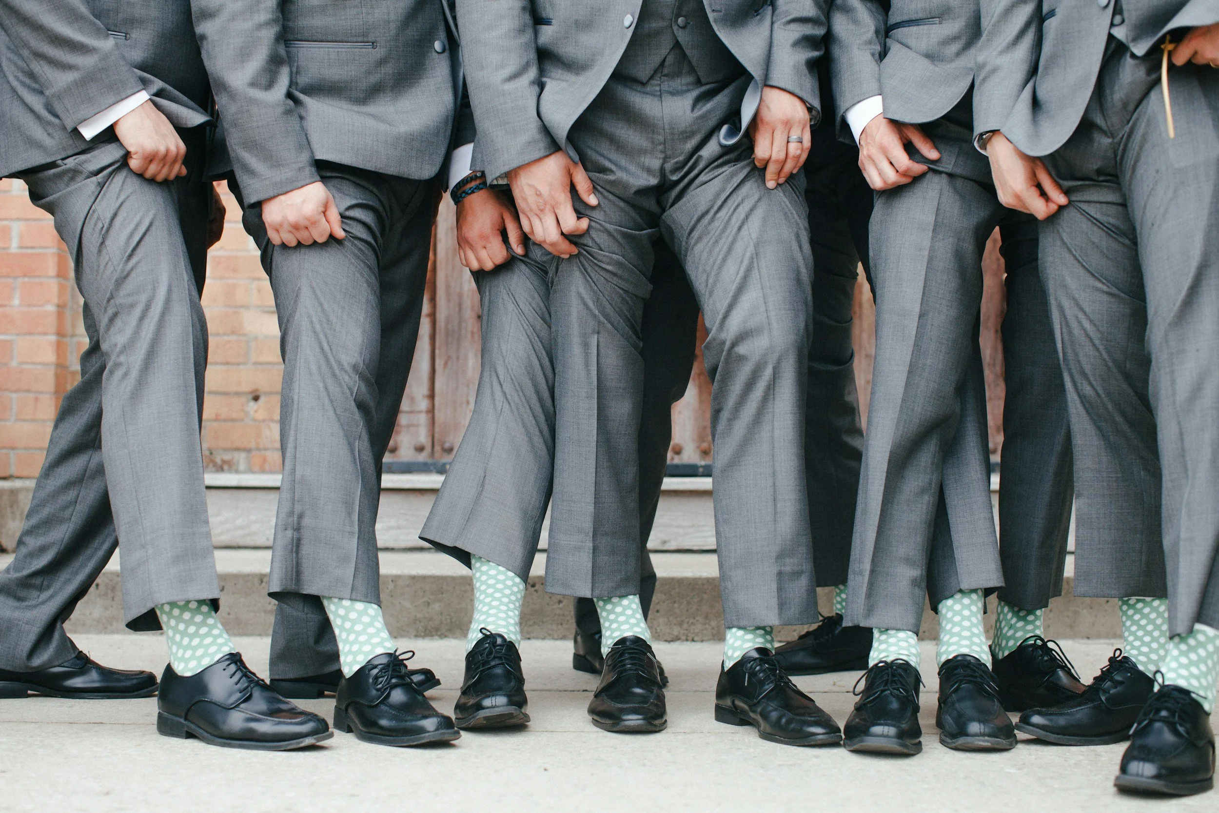 People in gray suits standing close together, with their hands and legs visible, wearing black shoes and green polka-dotted socks.