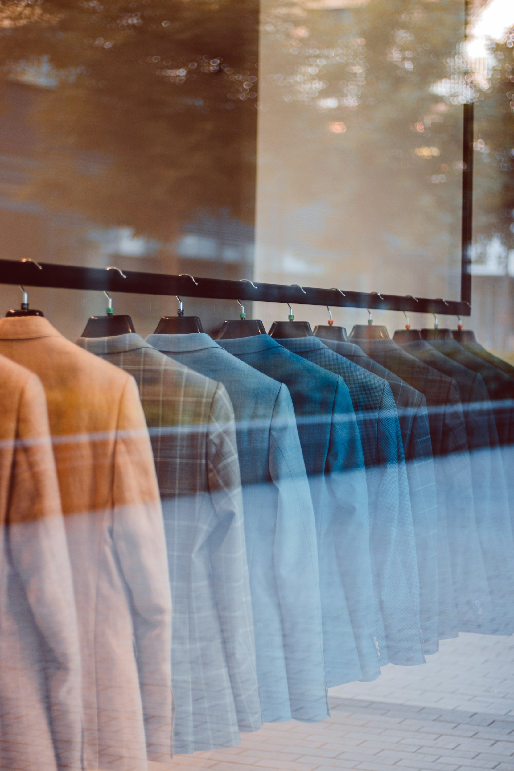 A row of men's suits in various colors hanging on a rack, viewed through a window with reflections of outside trees and buildings.