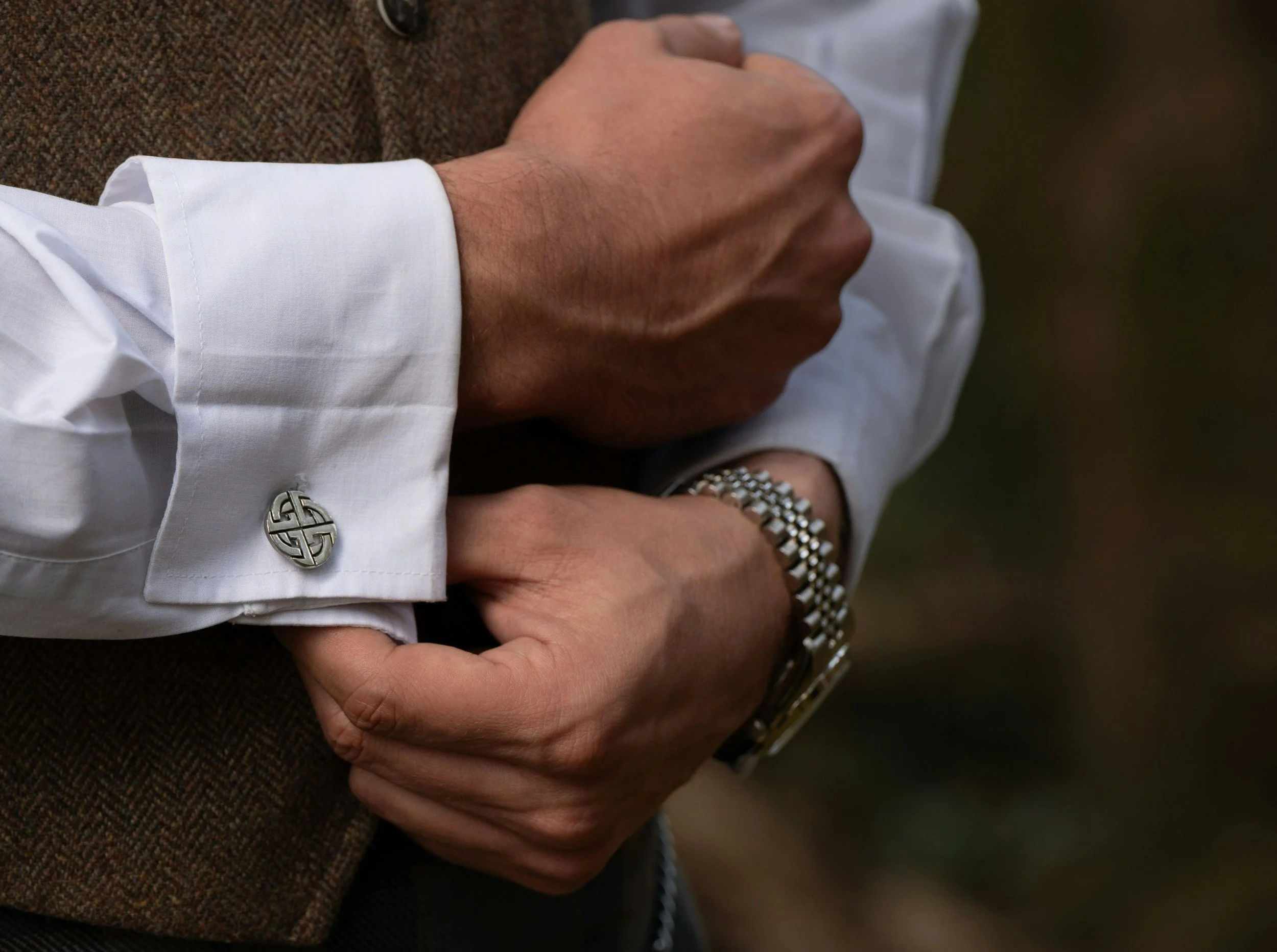 Close-up of a person wearing a white shirt with silver cufflinks, a brown textured jacket, and a silver watch, holding hands with another individual wearing a brown jacket.