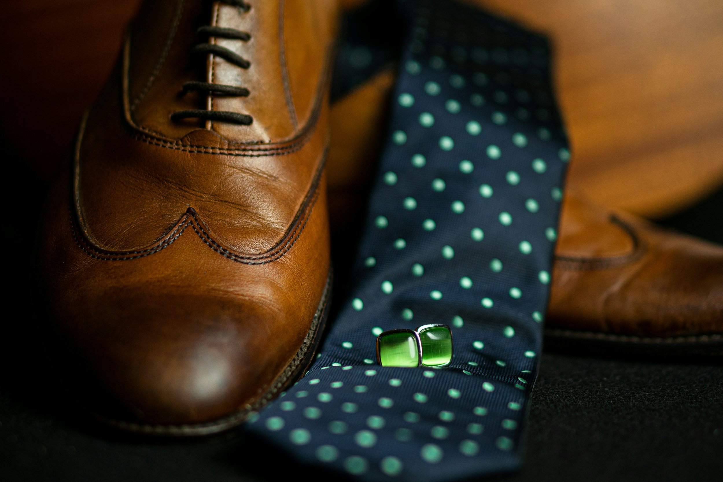 Close-up of a brown leather dress shoe, blue polka dot tie, and a pair of green glasses on a dark surface.