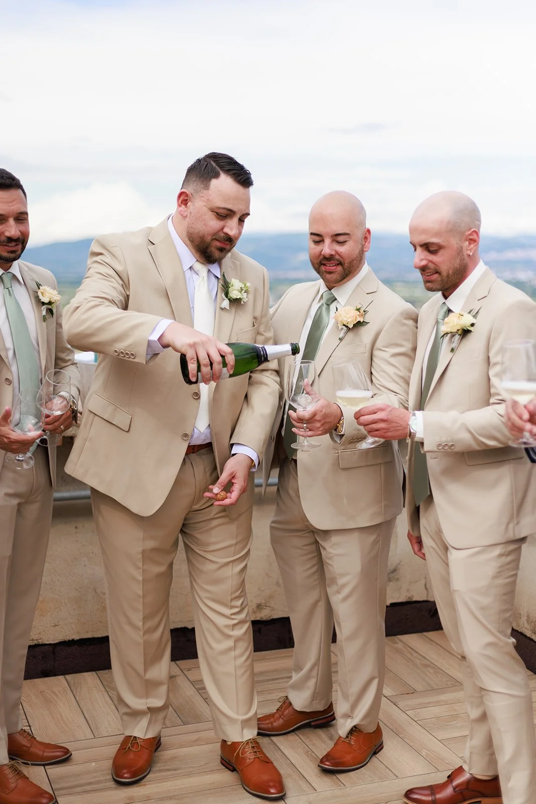 Groom and groomsmen in beige suits at outdoor wedding celebration, pouring champagne into glasses.