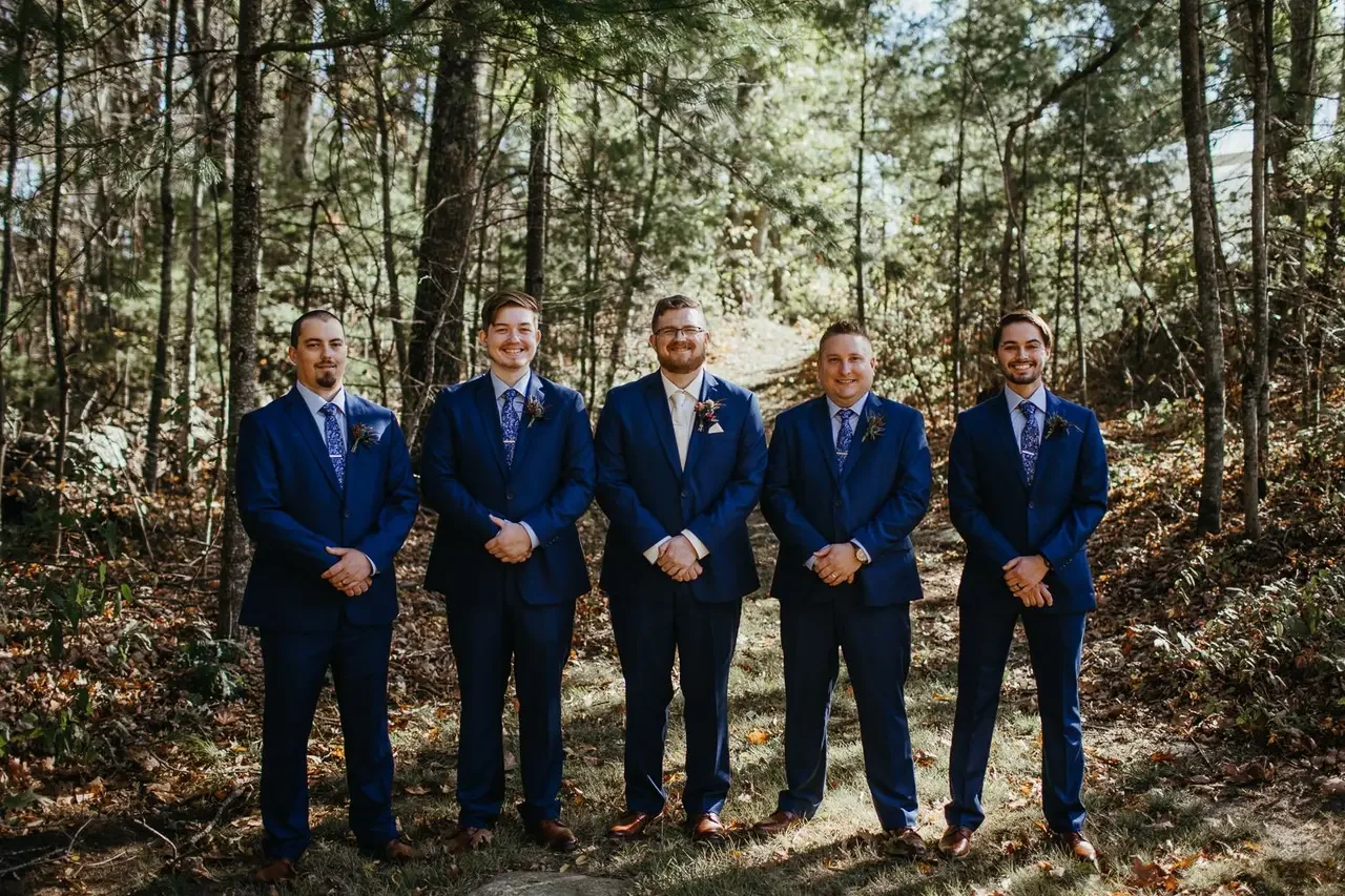 Group of five men dressed in matching blue suits standing outdoors in a wooded area.
