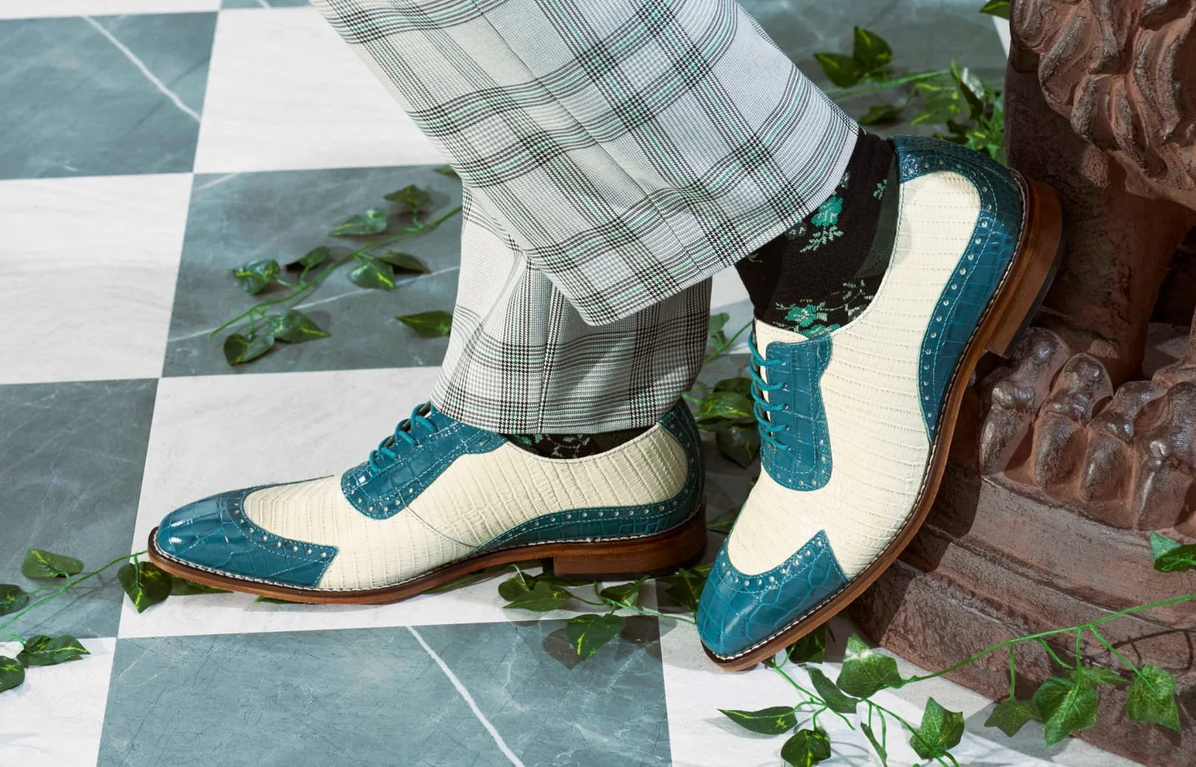 Close-up of a person's feet wearing beige and blue brogue shoes, black patterned socks, plaid pants, and standing on patterned tile floor with green plant leaves nearby.