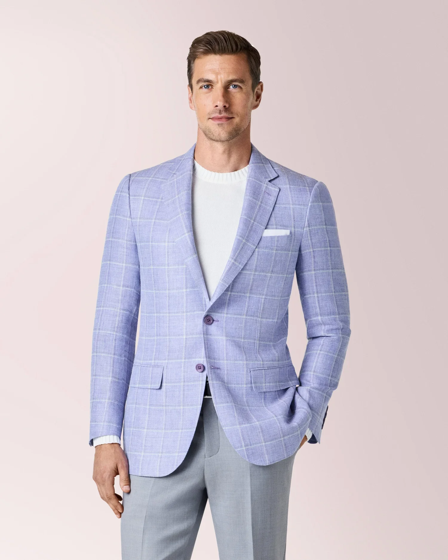 A man with short brown hair and blue eyes wearing a light blue checkered blazer over a white sweater, with light gray pants, standing against a plain light pink background.