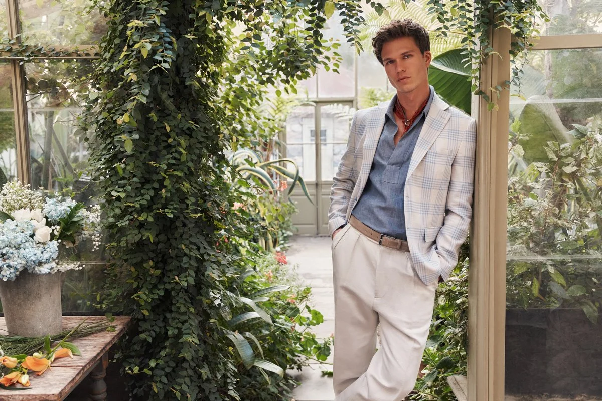 A young man with dark hair and fair skin leaning against a doorframe in a greenhouse, wearing a light-colored plaid blazer, a blue shirt, beige trousers, and a red necklace, surrounded by lush green plants and flowers.