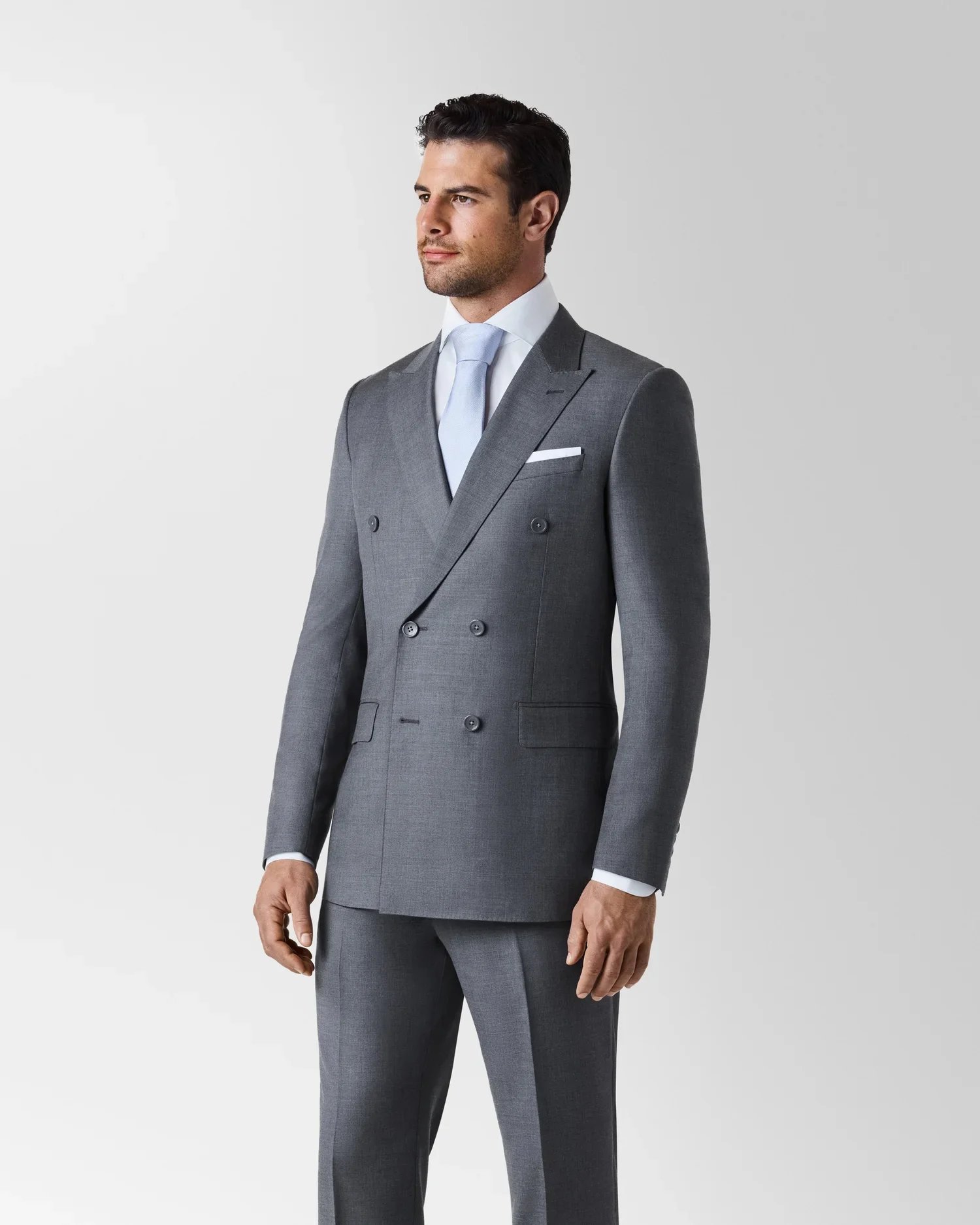 Man in a gray double-breasted suit with a light blue tie standing against a plain background.