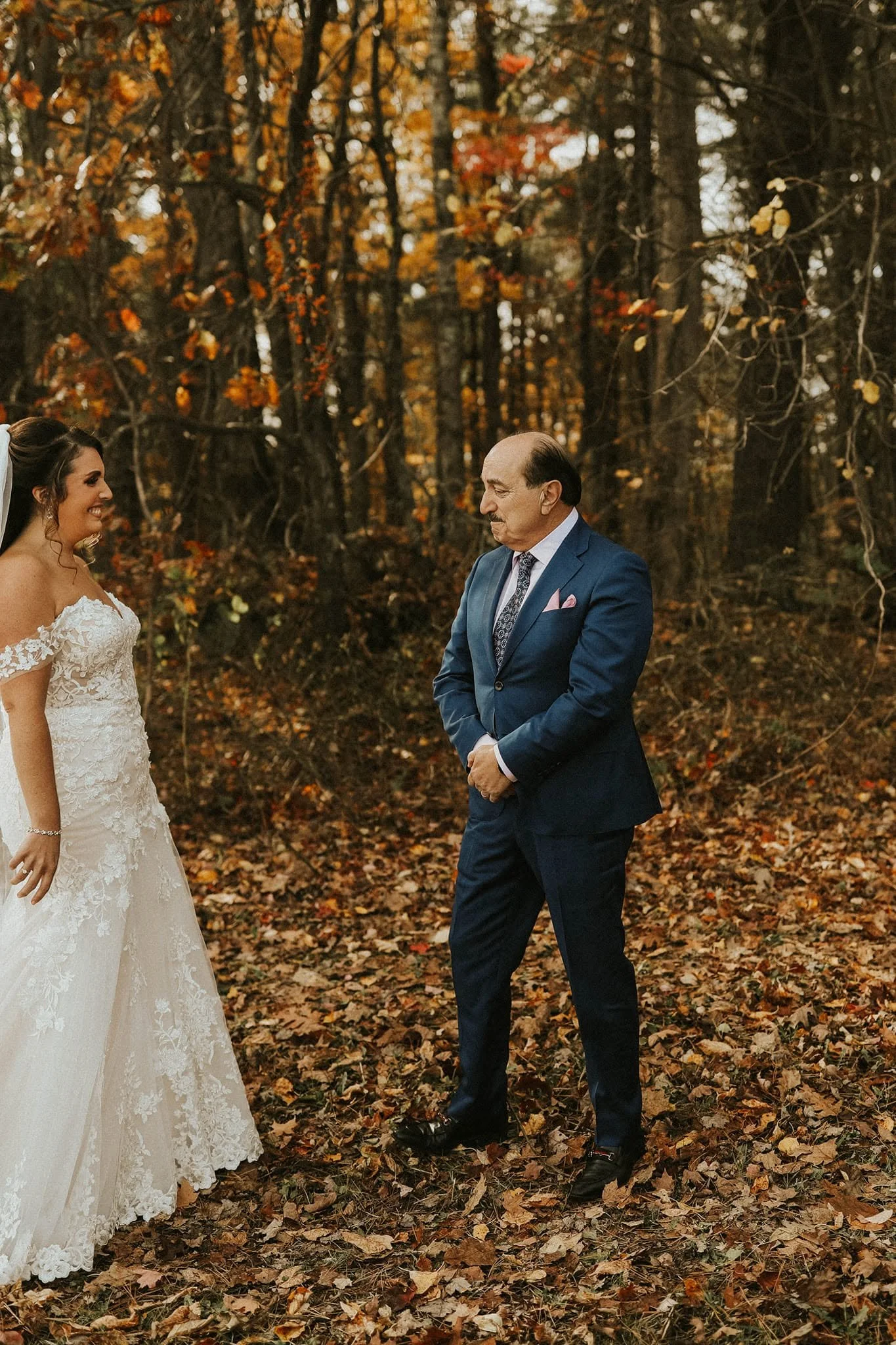 A bride in a white lace wedding gown smiling and looking at a man in a blue suit, standing in a wooded area with fallen autumn leaves.