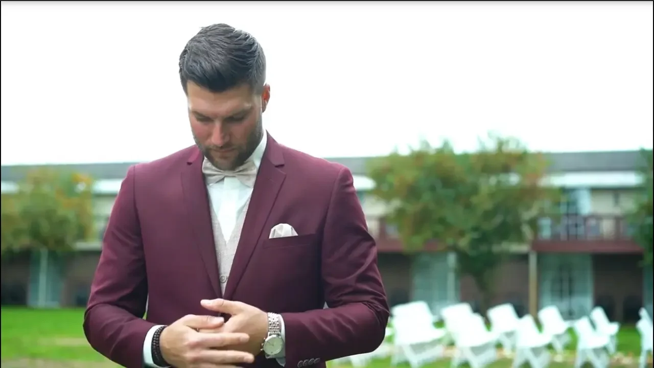 A man in a maroon suit and white shirt adjusting his jacket outdoors, with chairs and a building in the background.