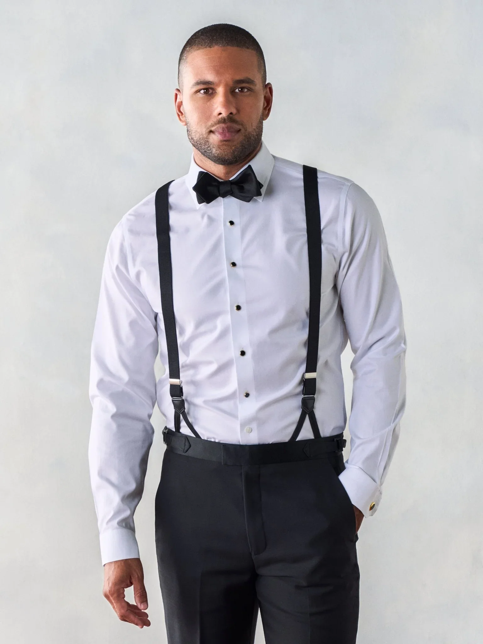 A man wearing a white dress shirt with black suspenders, a black bow tie, and black dress pants. He is standing against a plain background, with one hand relaxed by his side.