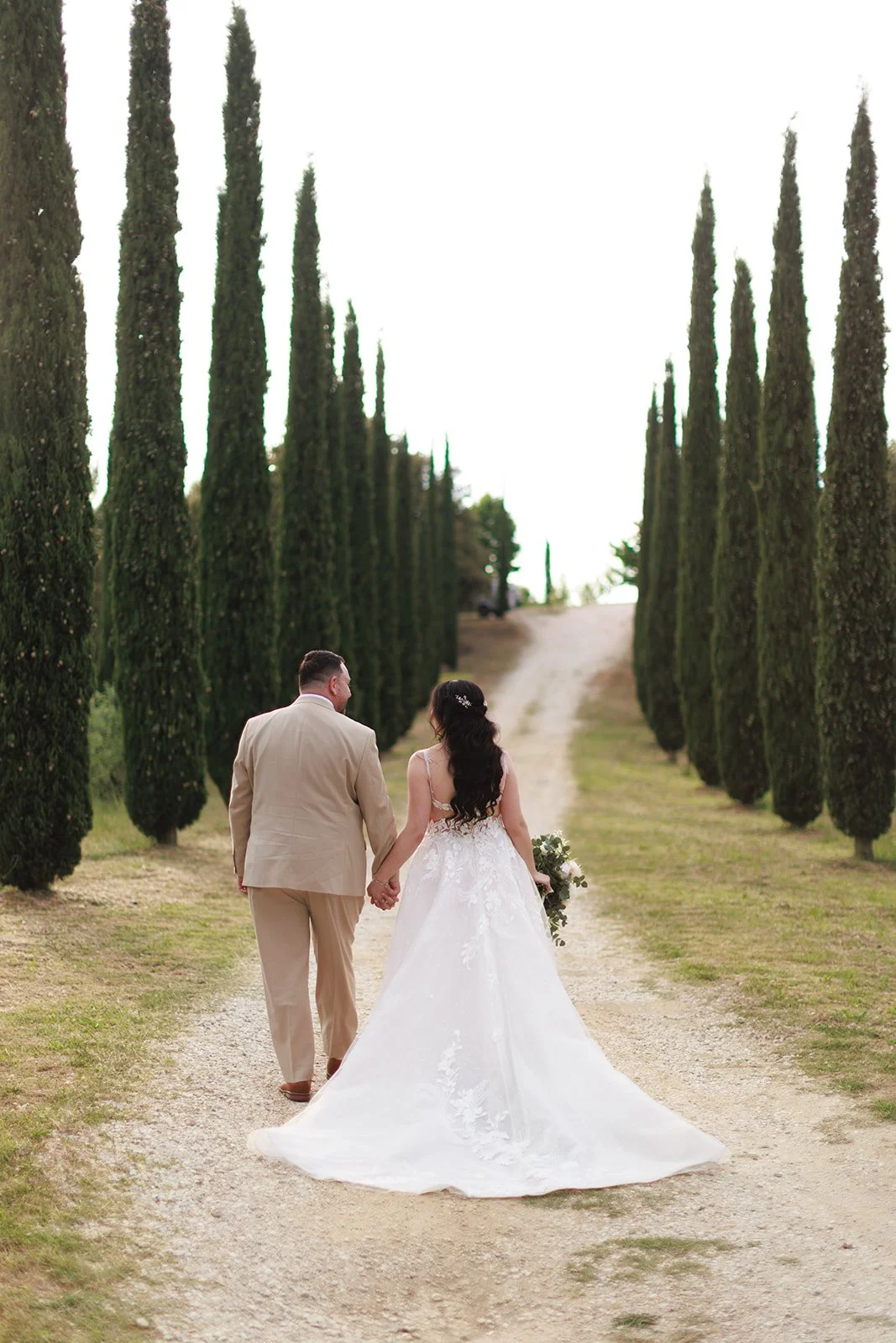 A bride and groom walking hand in hand along a path lined with tall cypress trees during sunset, with the bride in a white wedding gown and the groom in a beige suit.