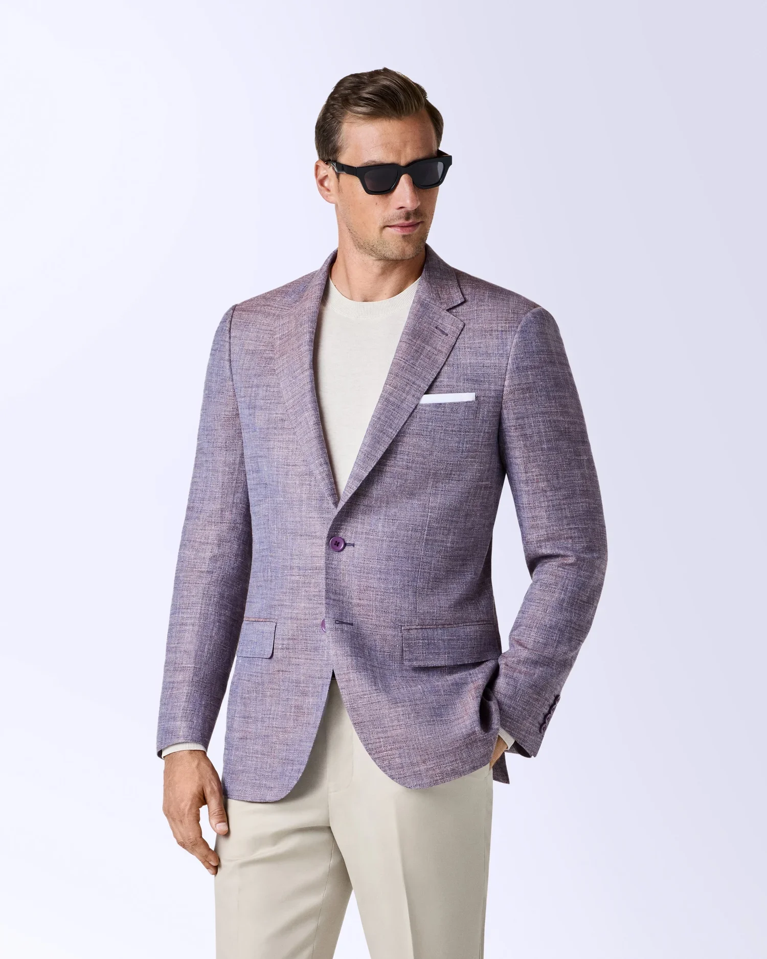 A man wearing dark sunglasses, a light purple plaid blazer, a white shirt, and beige pants standing against a plain white background.