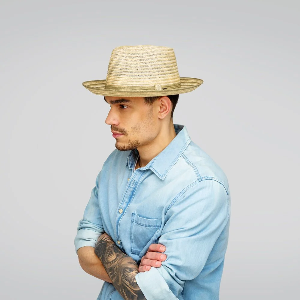 A young man in a light blue denim shirt with rolled-up sleeves, wearing a wide-brimmed straw hat, standing with crossed arms and looking to the side against a plain light gray background.