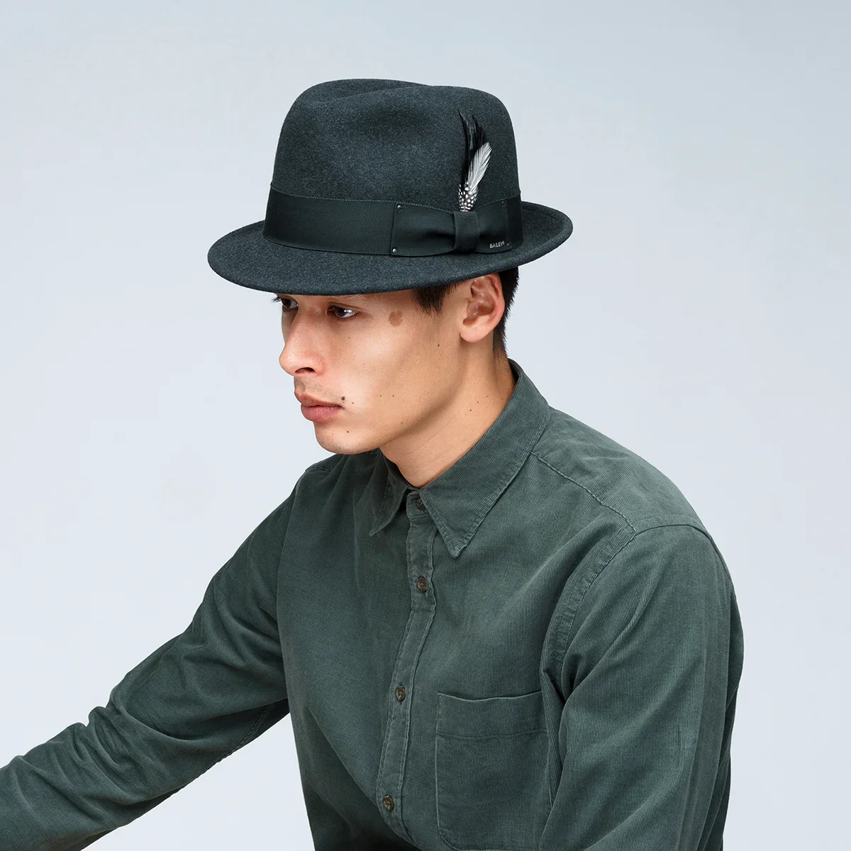 A young man wearing a dark gray felt fedora hat with a black band and decorative feathers, dressed in a dark green button-up shirt, against a plain light background.