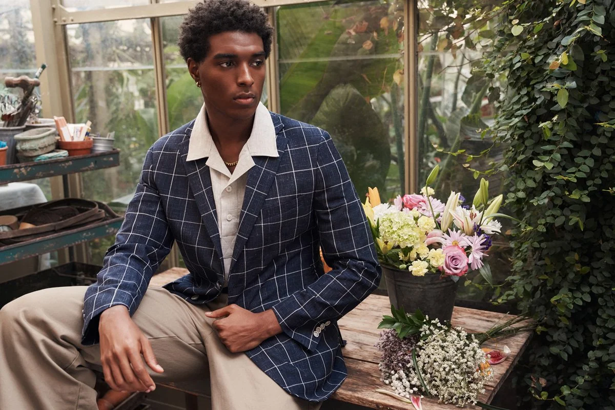 A young man with dark skin and short curly hair, dressed in a beige shirt and a checkered navy blazer, sits on a wooden table in a greenhouse or botanical garden setting. Next to him is a large pot of mixed pink and white flowers, surrounded by green