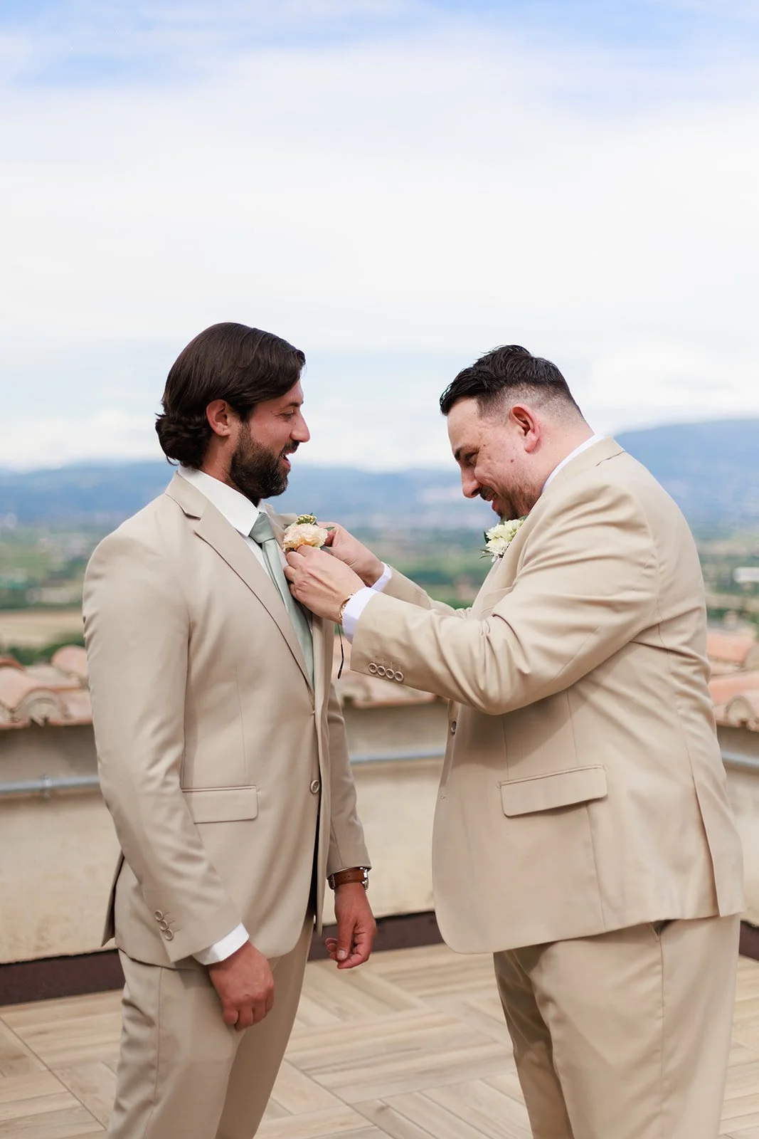 Two men in beige suits at a wedding, one is pinning a boutonniere on the other's lapel on a rooftop with a scenic landscape in the background.