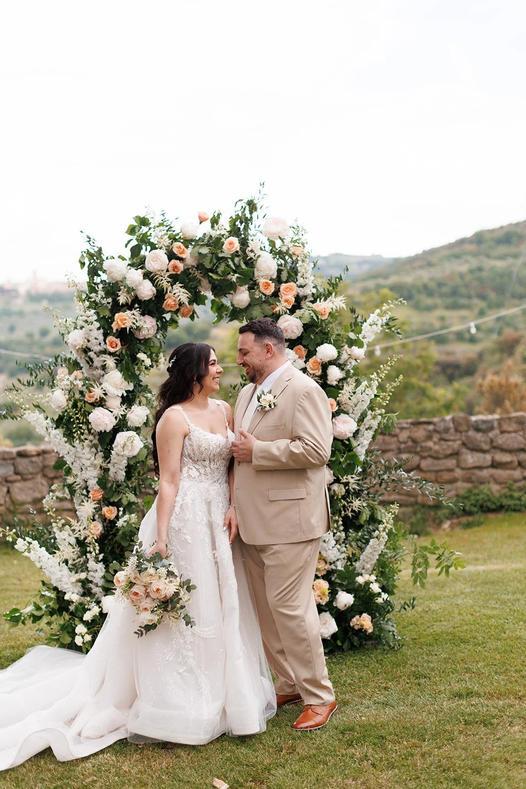 A bride and groom standing close together during their wedding ceremony, smiling at each other, with a large floral arch behind them outdoors on a grassy area.