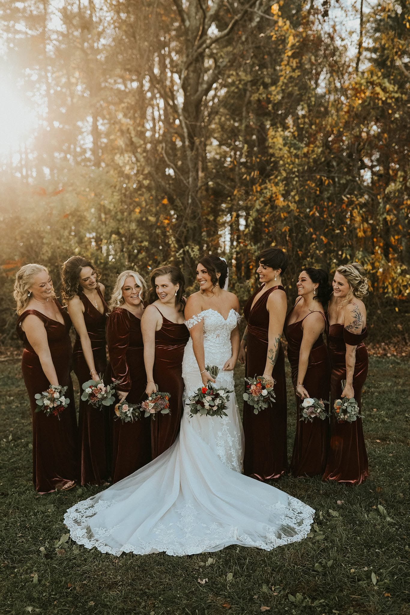 A bride and seven women, all wearing burgundy dresses, standing outdoors in a park with autumn foliage, smiling and holding bouquets of flowers.