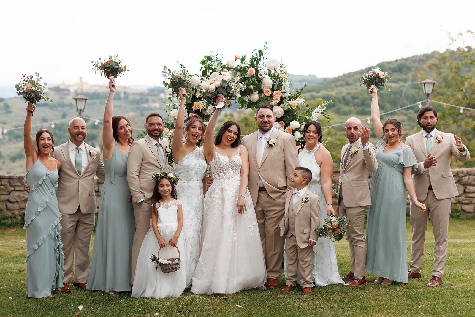 A wedding party outdoors, including the bride and groom, bridesmaids, groomsmen, children, and family, celebrating with smiles and raising bouquets in front of a floral arch and scenic hills.