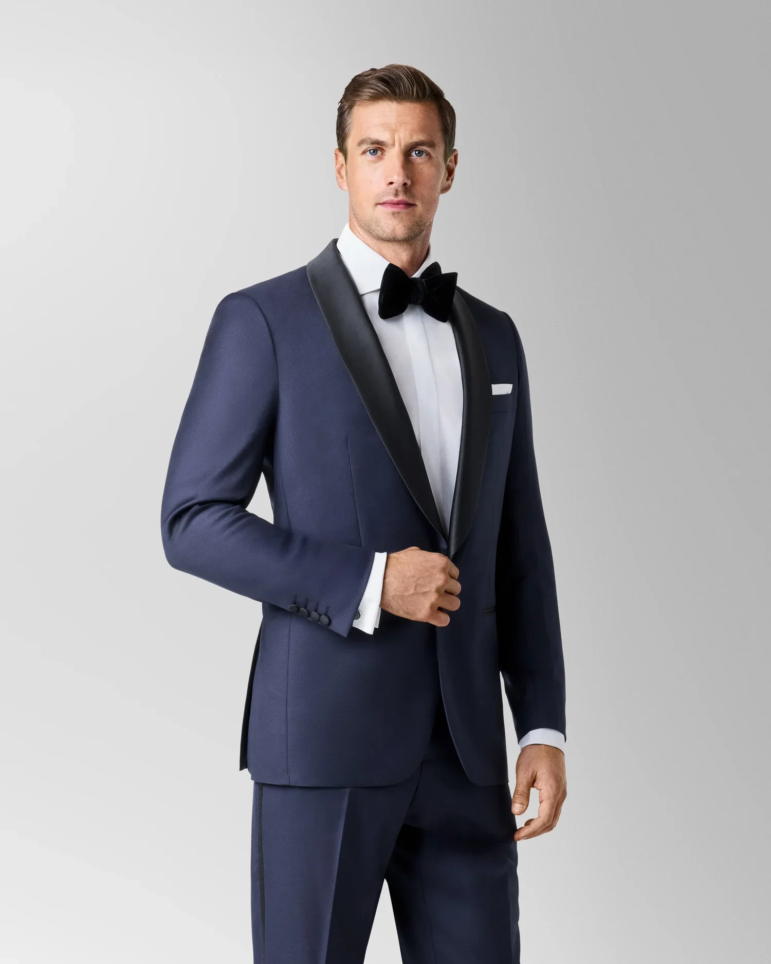 Man in a navy tuxedo with a white shirt and black bow tie standing against a plain gray background.