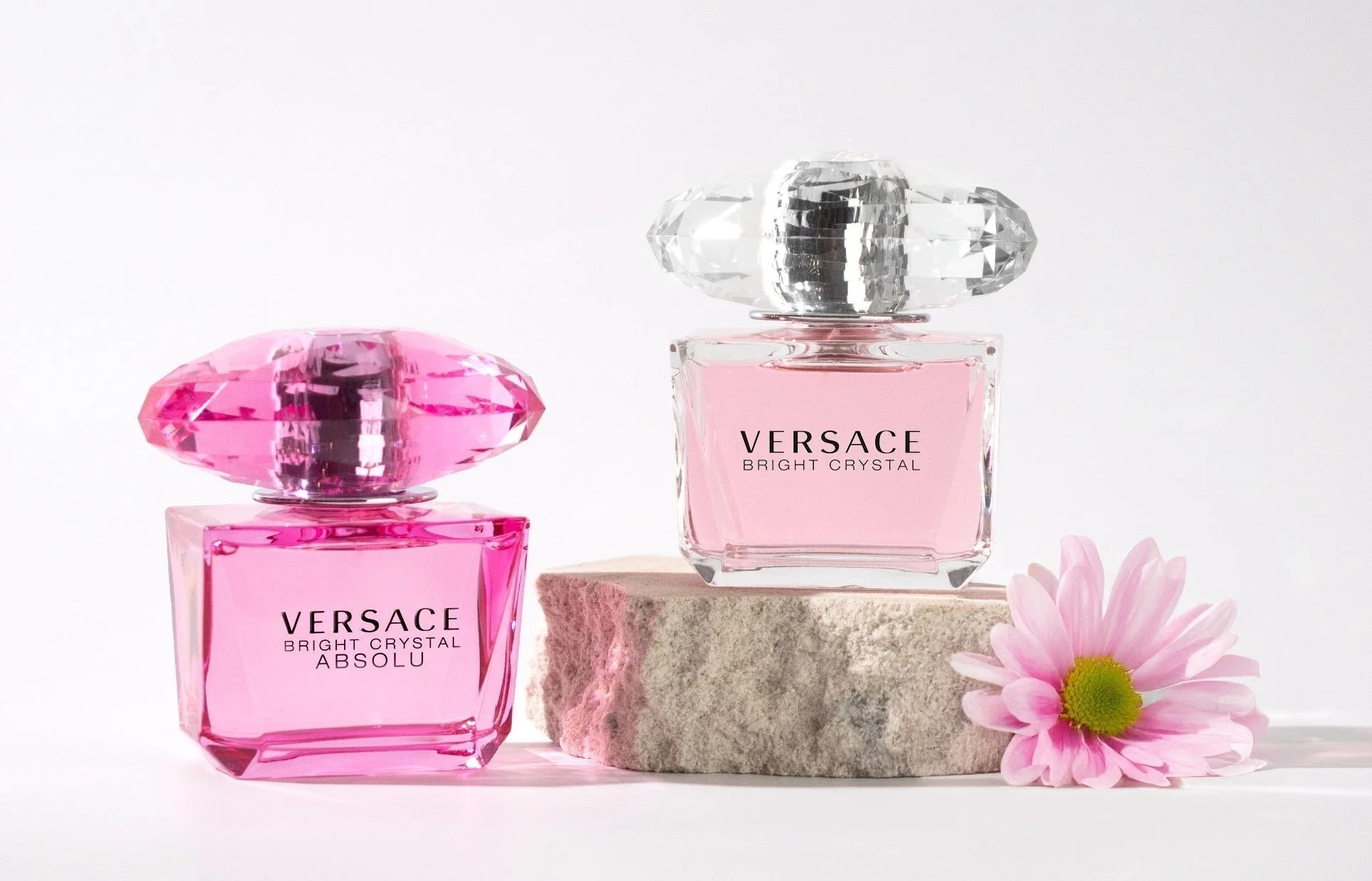 Two Versace Bright Crystal perfume bottles with pink liquid, one labeled Absolu and the other labeled Bright Crystal. The bottles have diamond-shaped caps, and there is a pink flower with yellow center beside a round stone platform on a light-colored