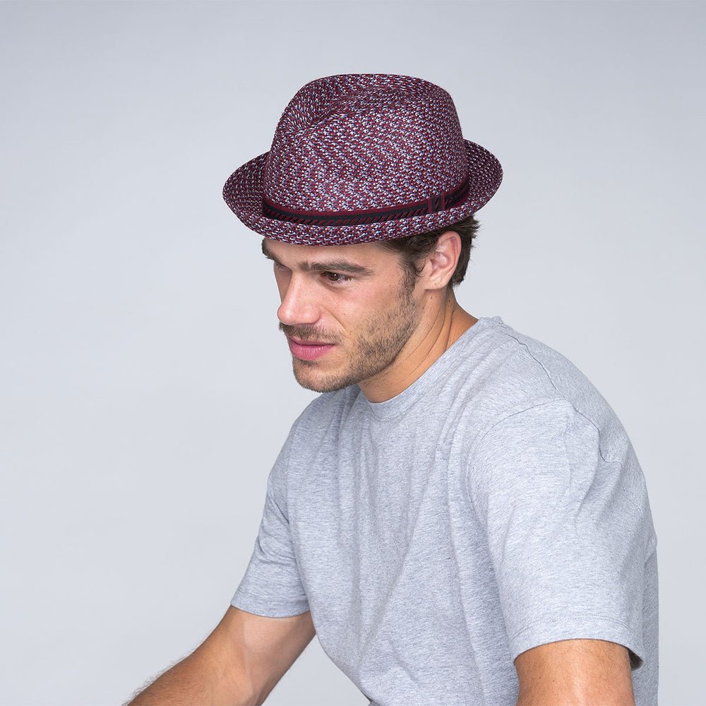 Man wearing a multicolored fedora hat and a light gray t-shirt, looking downward with a slight smile, against a plain light background.