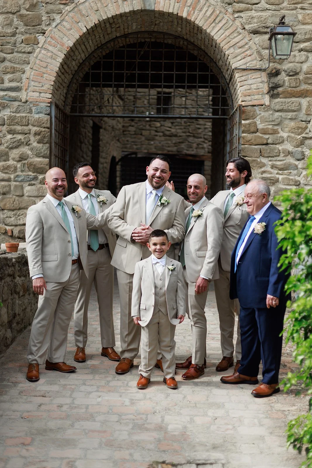 A groom and a young boy, possibly a ring bearer, surrounded by six groomsmen and an older man, all dressed in beige suits and standing under a stone archway in an outdoor setting