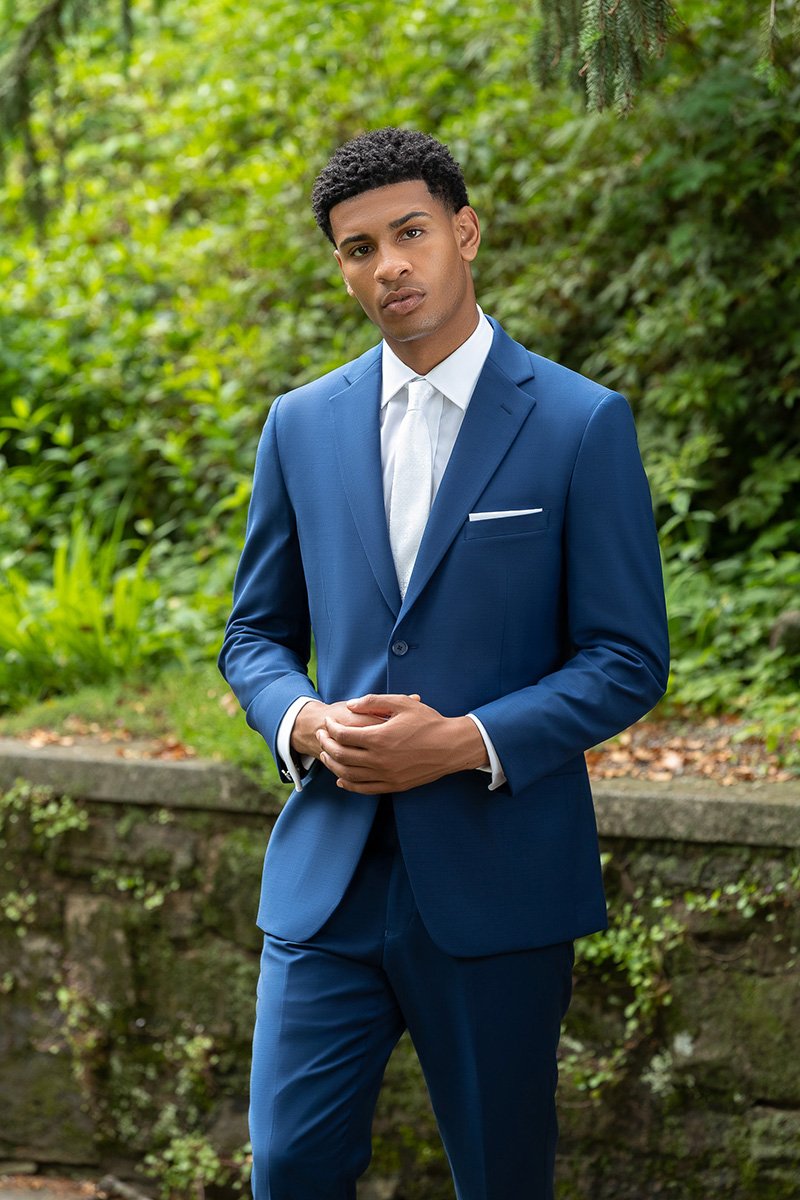 Young man in a blue suit standing outdoors with greenery in the background.