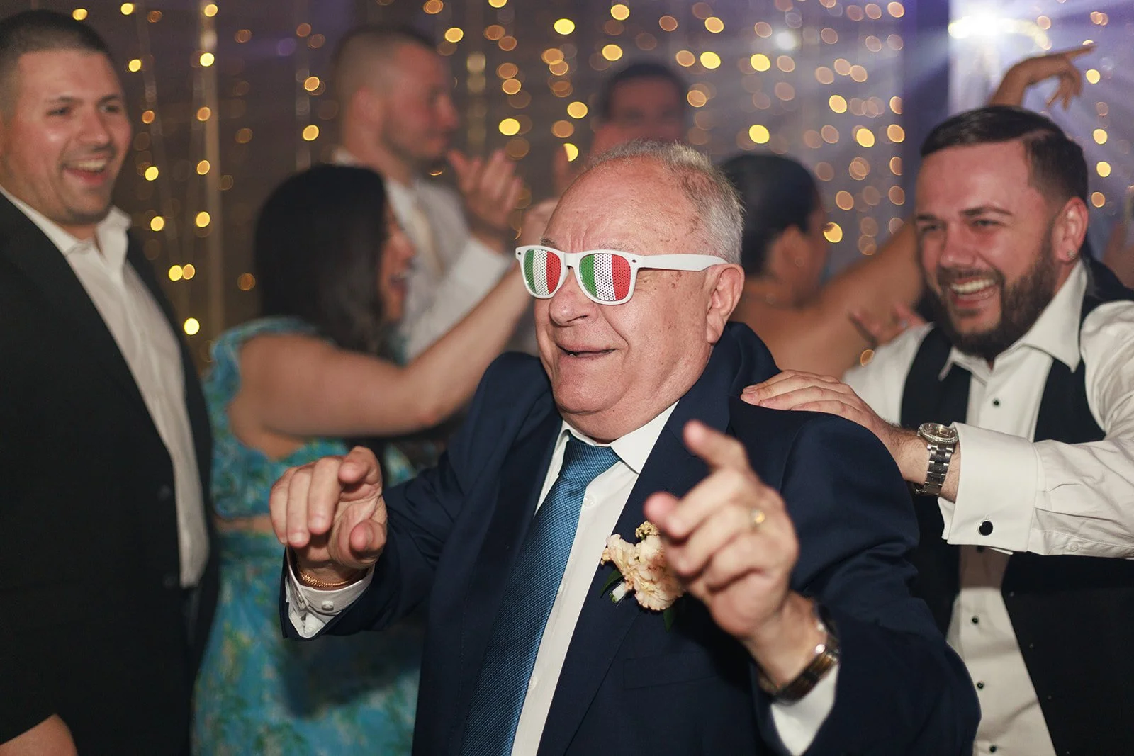 A group of people celebrating at a party, with a man in the center wearing Italian flag sunglasses, dancing and smiling, surrounded by friends in formal attire, with festive lights in the background.