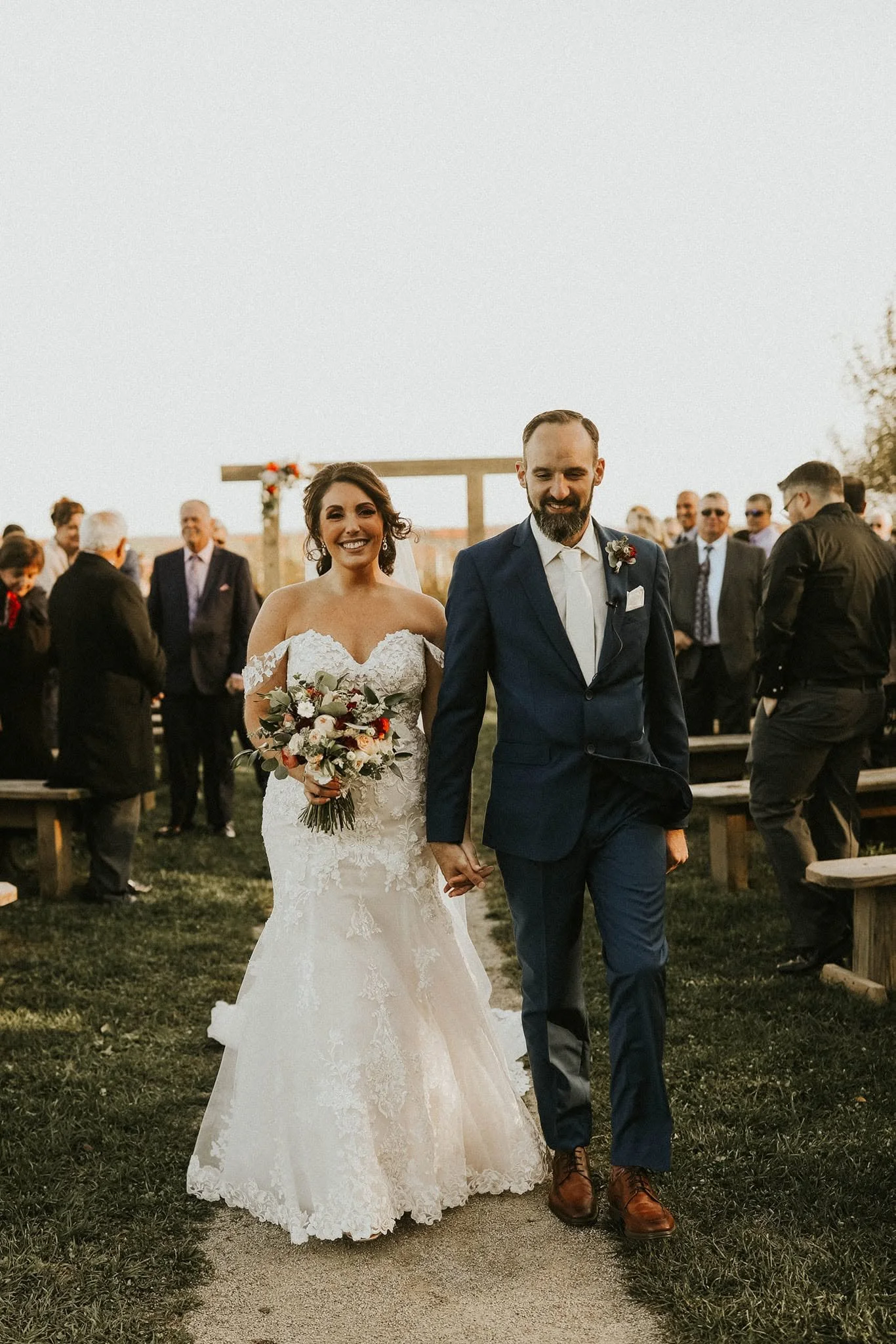A newly married couple holding hands and walking outside during their wedding ceremony, surrounded by guests, with a wooden arch and floral decorations in the background.