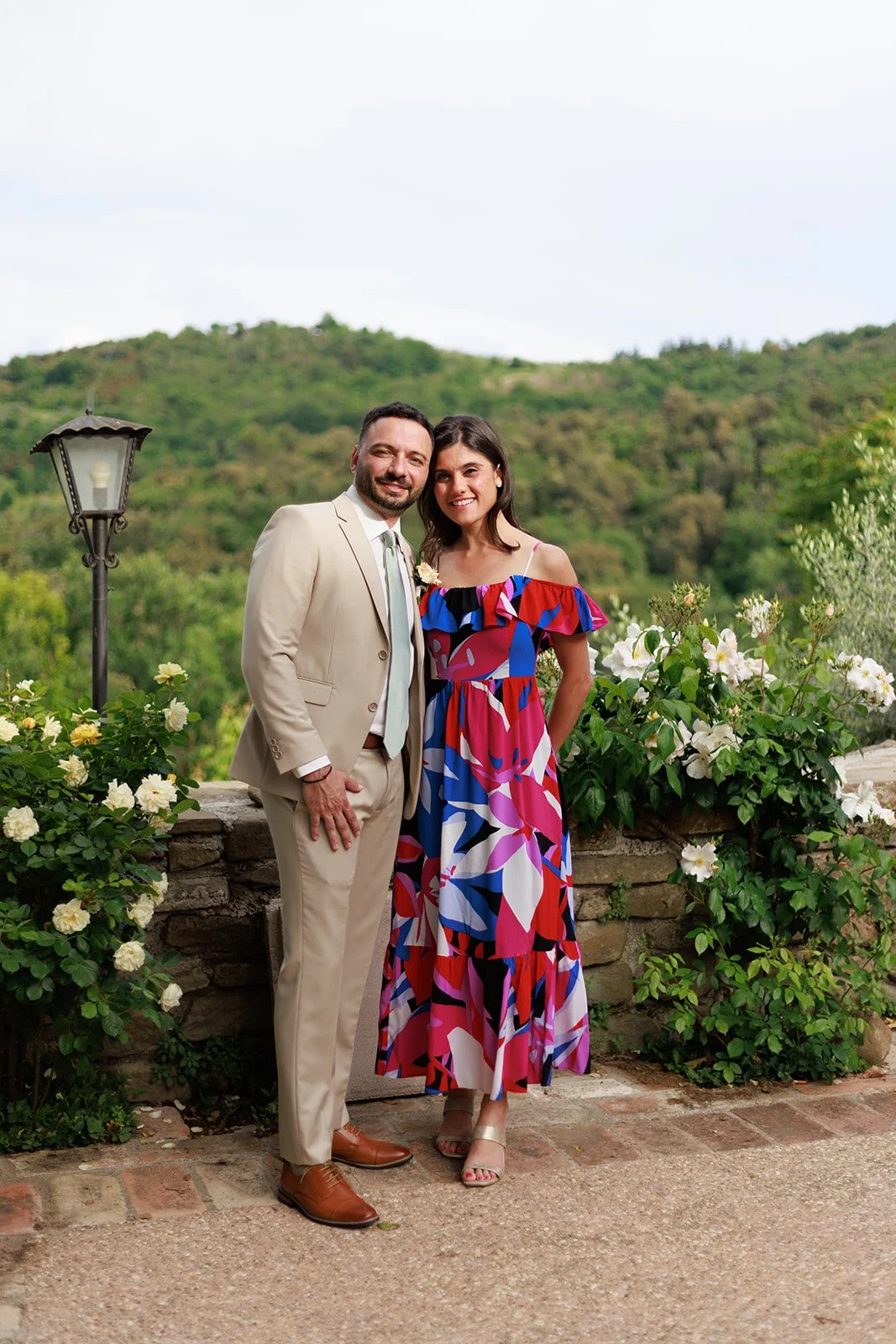 A man and woman standing outdoors on a sunny day, smiling and posing for a photo, with greenery and hills in the background, surrounded by white flowers and a vintage street lamp.