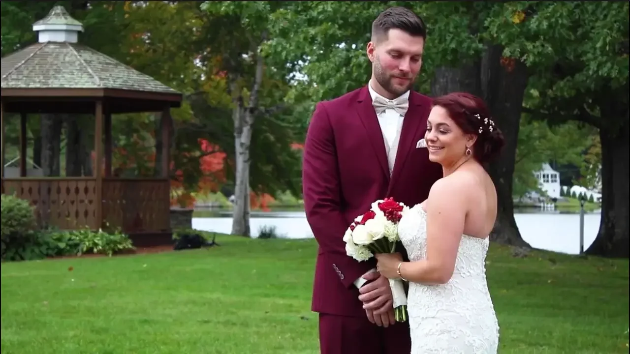A bride and groom stand together outdoors, holding a bouquet of white and red flowers, with trees and a small wooden gazebo in the background.