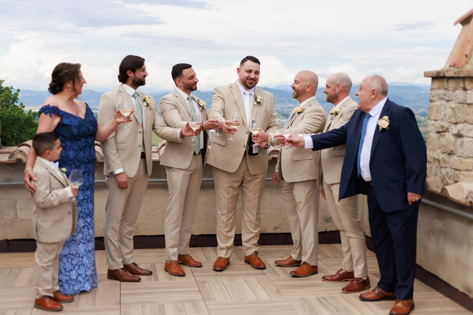 A group of people at a wedding celebration, including a bride and groom among seven men dressed in beige suits, a woman in a blue dress, and a young boy in a beige suit, all holding champagne glasses and toasting outdoors with scenic mountains in the