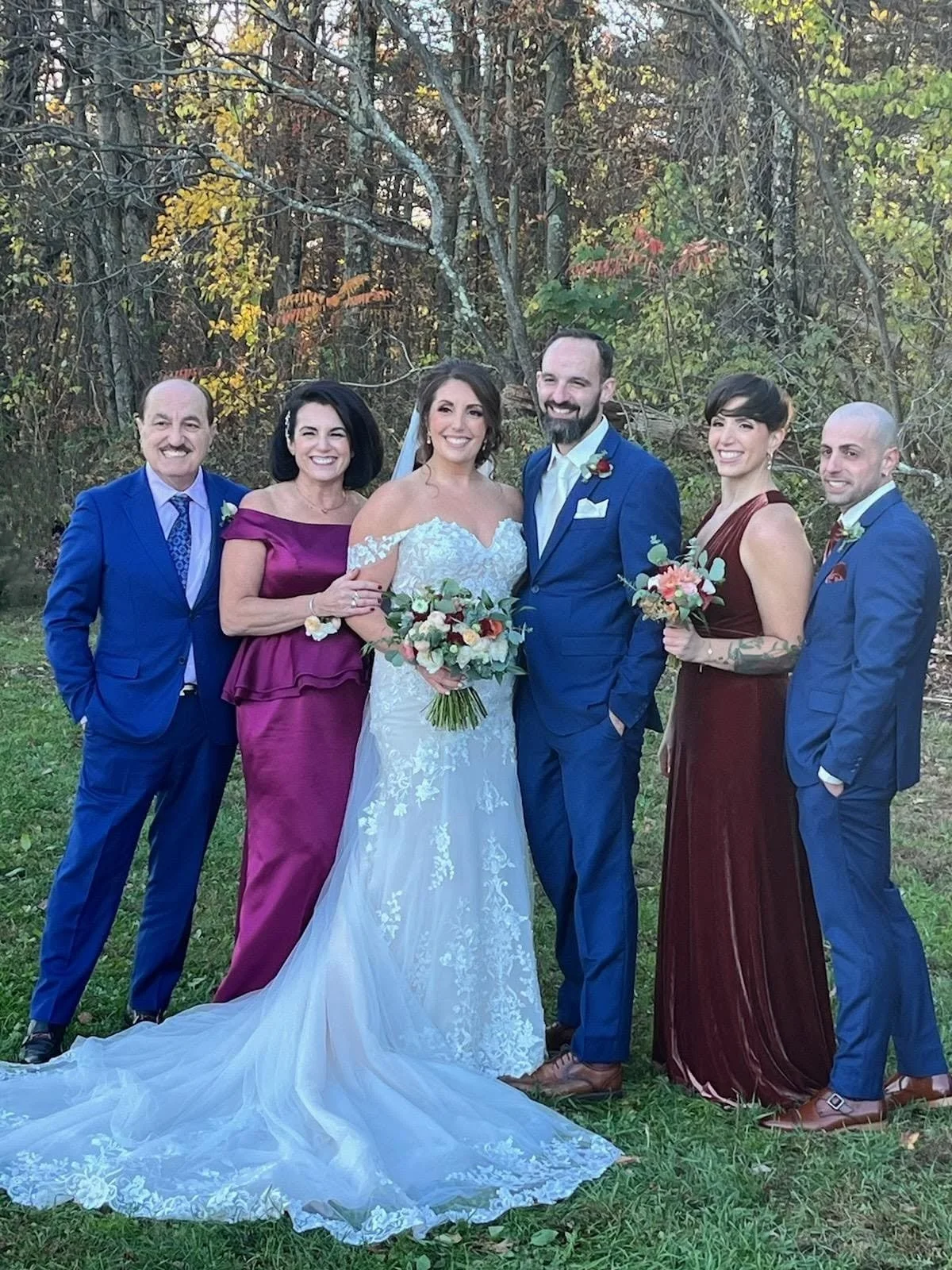 A group of seven people at a wedding, standing outdoors in front of trees. The bride is in the center wearing a white gown and holding a bouquet. The groom is to her right in a blue suit. Two women and two men are on either side, dressed in formal at