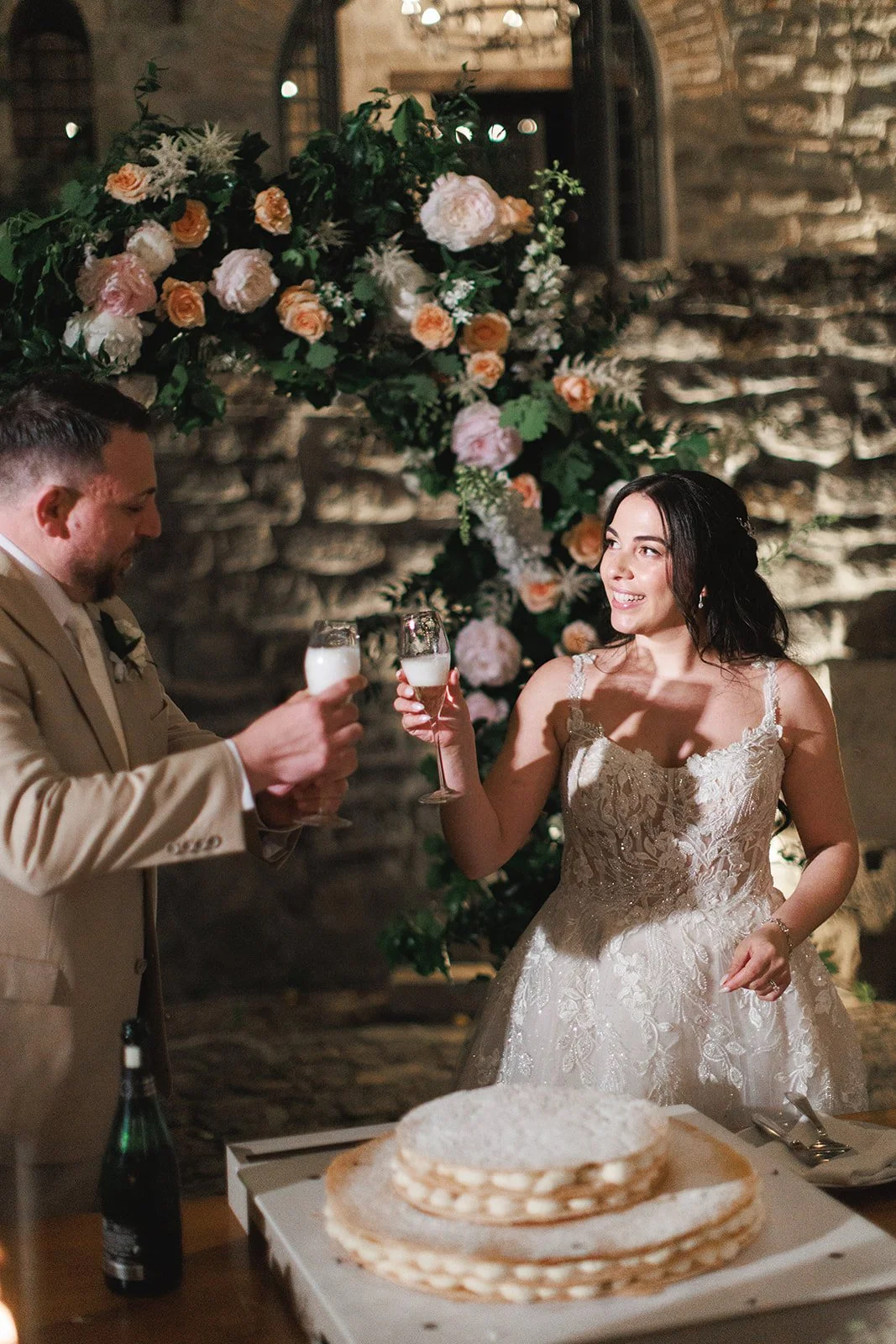 Bride and groom toasting with champagne at their wedding reception, with a floral backdrop and a layered wedding cake on the table.