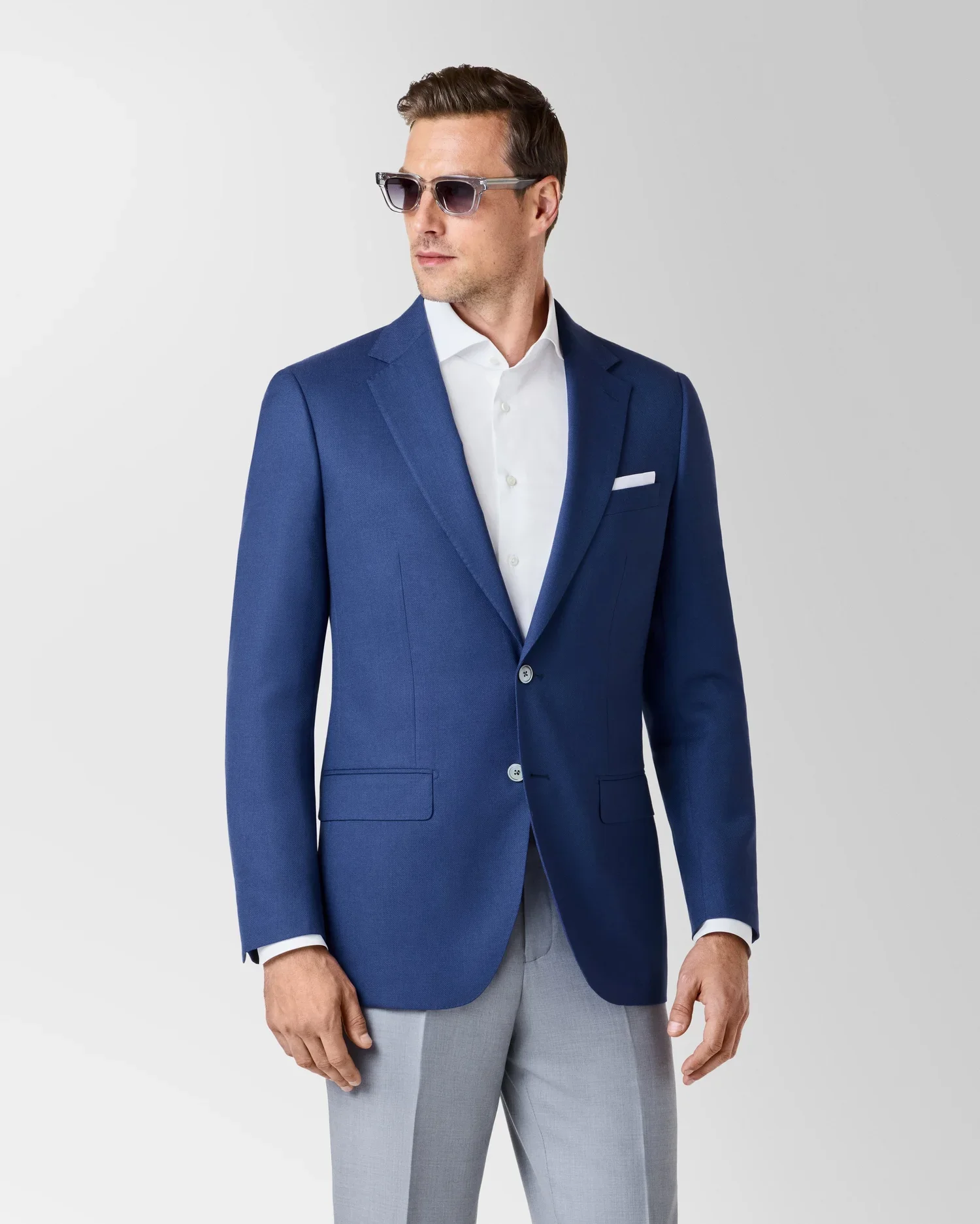 A man wearing a blue blazer, white dress shirt, light gray pants, black sunglasses, standing against a light gray background.