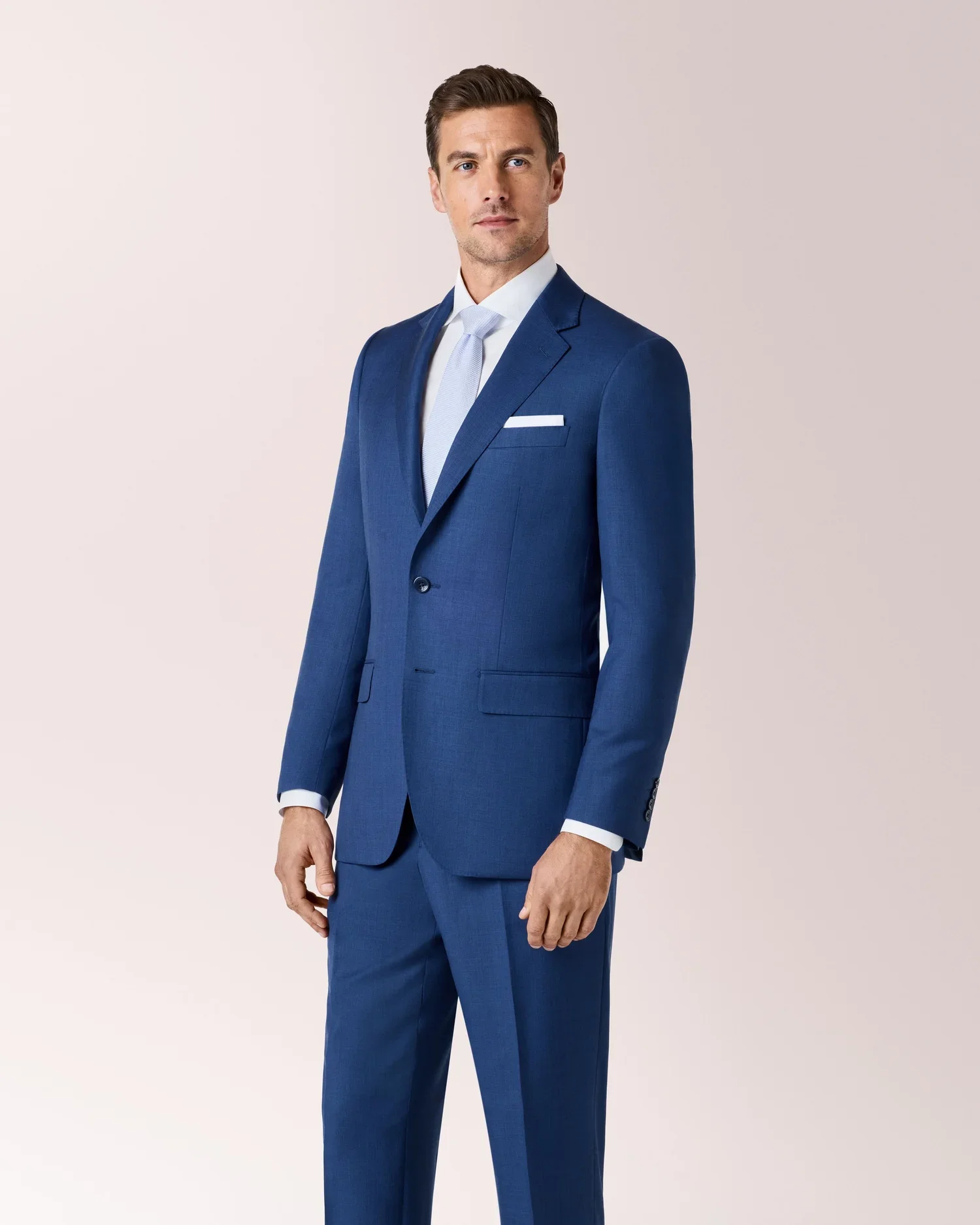 Man in a blue suit and light blue tie standing against a plain background.