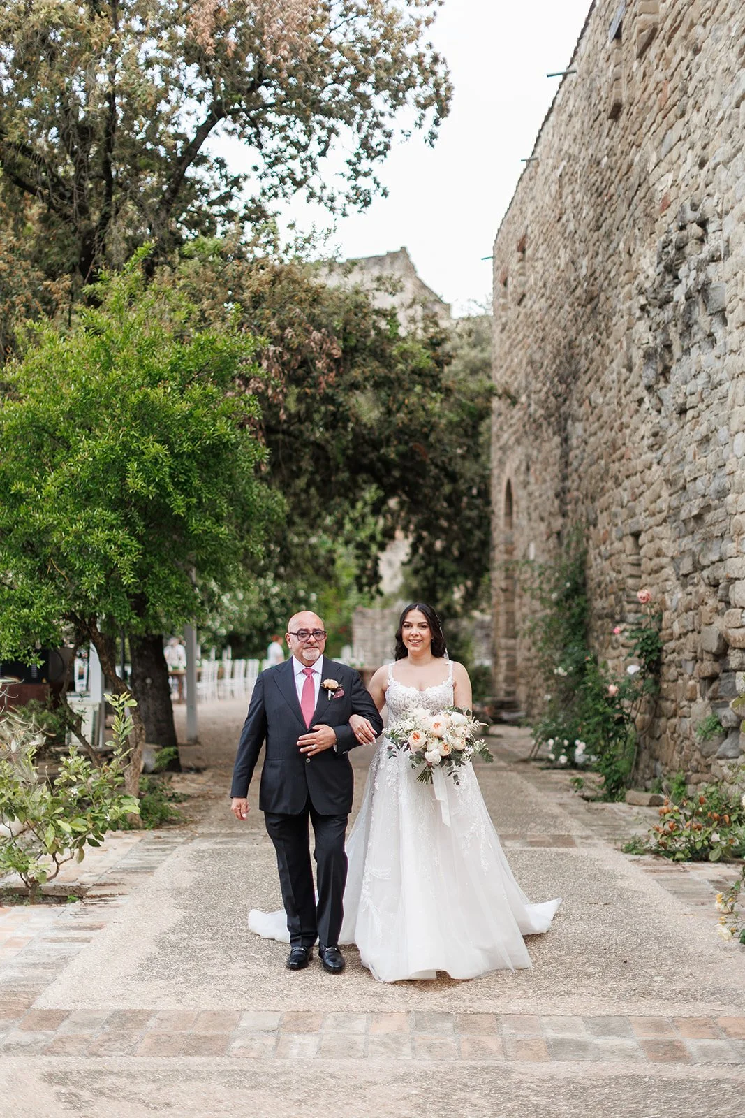 A bride in a white wedding dress holding a bouquet walking with an older man in a suit down a stone pathway lined with trees and flowers, next to a historic stone wall.