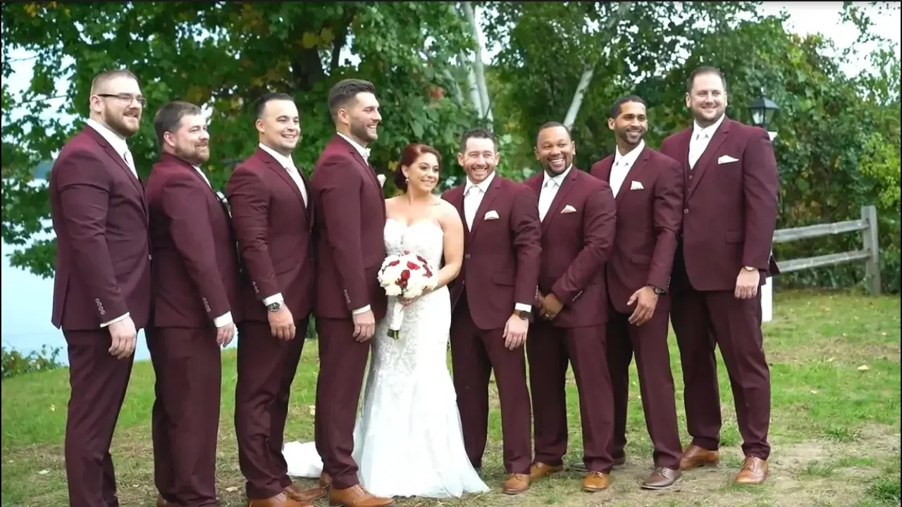 A wedding group photo shows a bride in a white wedding gown holding a bouquet, surrounded by nine groomsmen in matching maroon suits, outdoors with green trees and a wooden fence in the background.