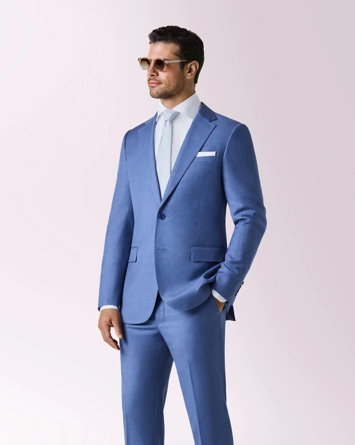 A man wearing a blue suit, white shirt, gray tie, and sunglasses, standing against a plain, light-colored background.