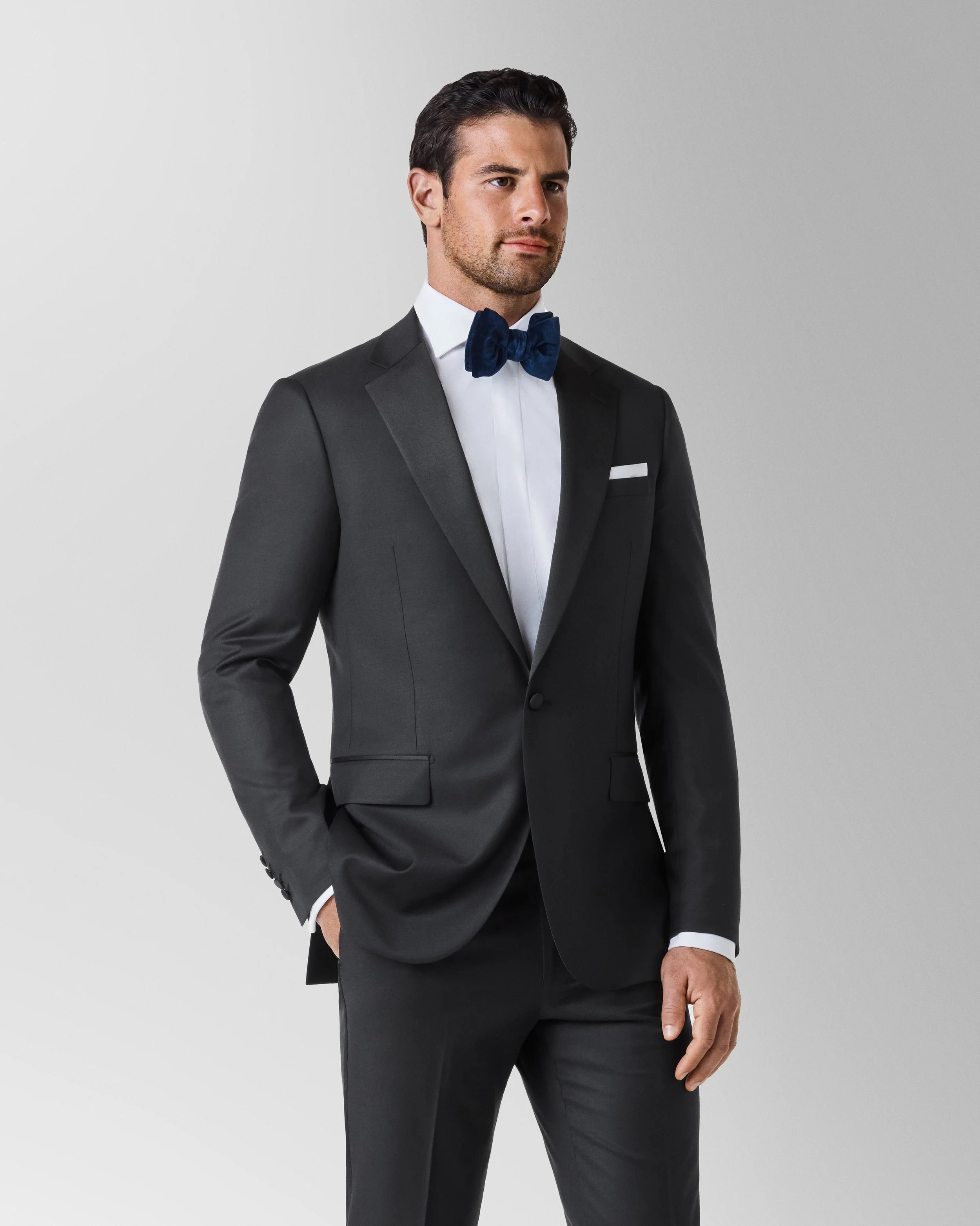 A man wearing a black tuxedo with a white shirt and a navy blue bow tie posing against a plain light gray background.