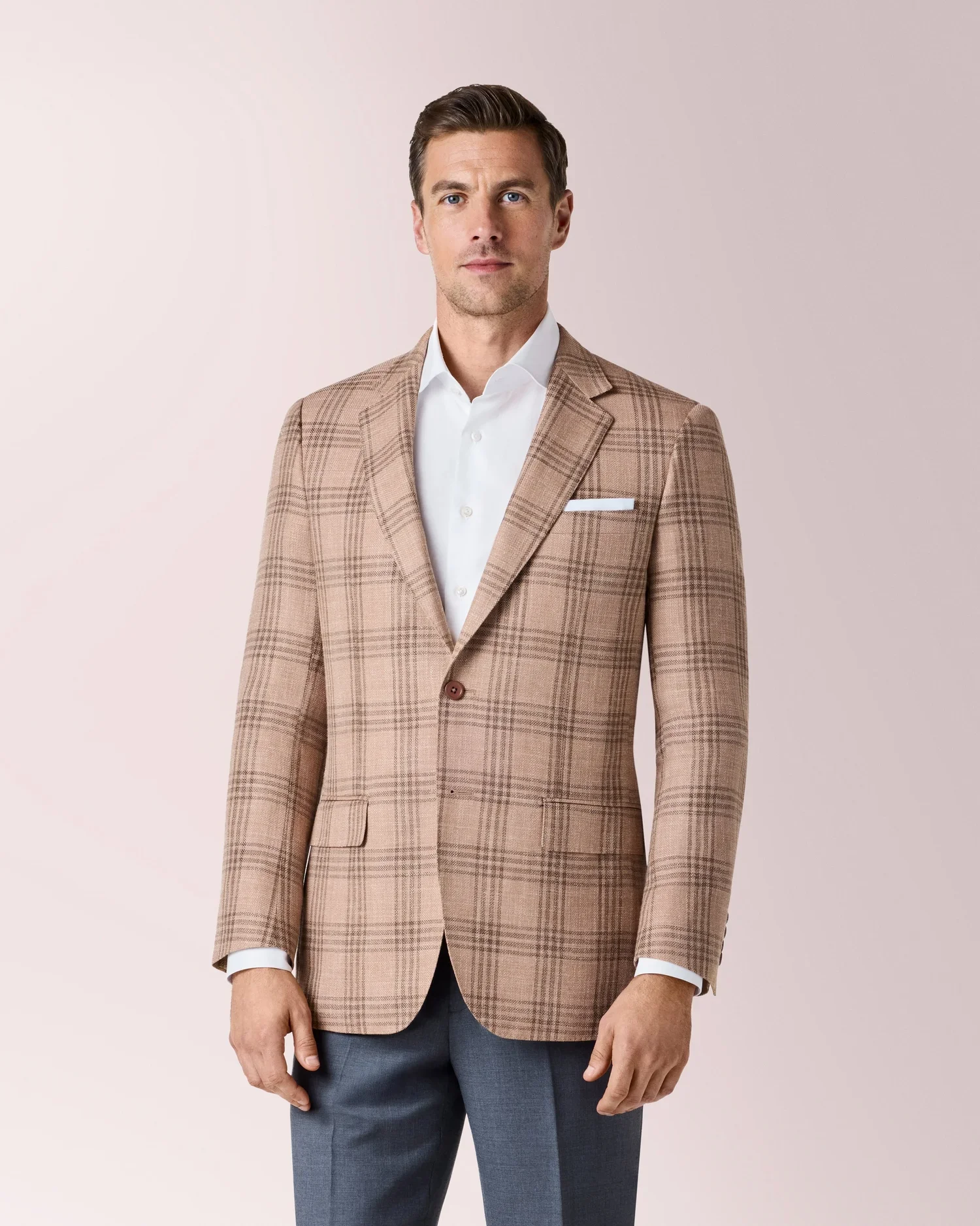 A man wearing a beige plaid blazer and light blue pants, standing against a light pink background.