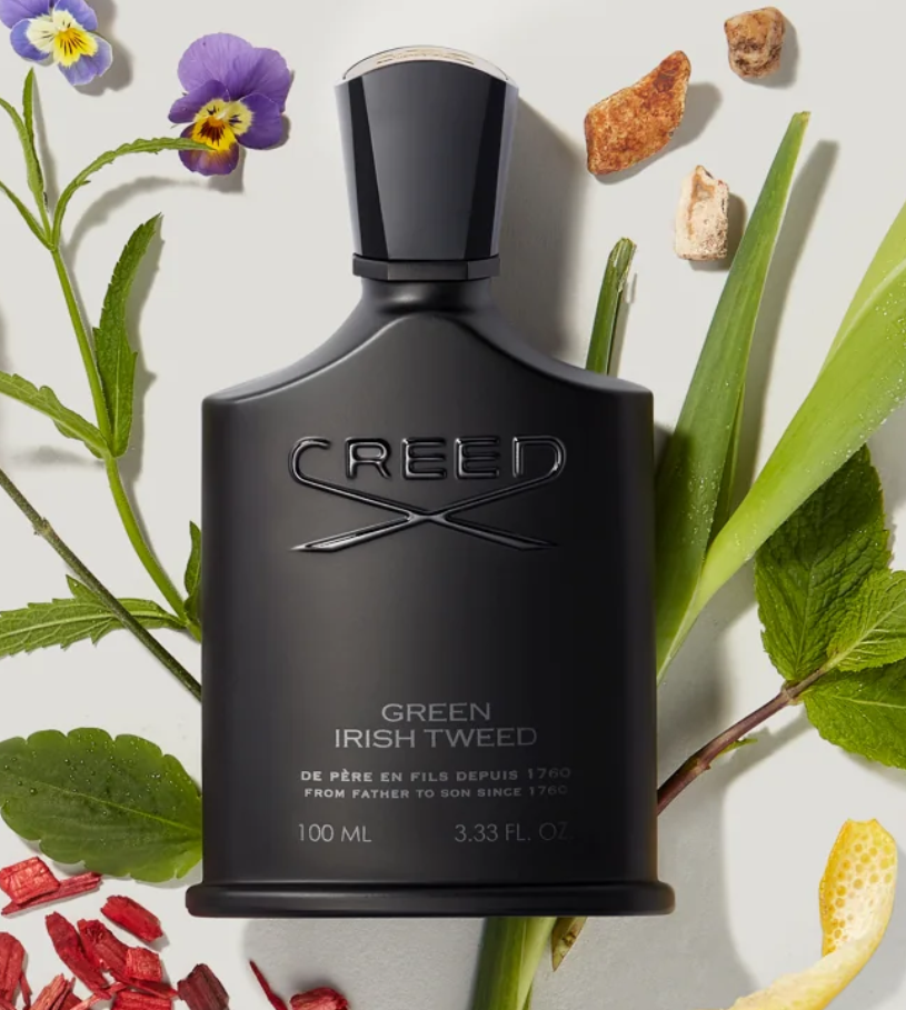 A black bottle of Creed Green Irish Tweed cologne surrounded by greenery, purple and yellow flowers, and small stones.