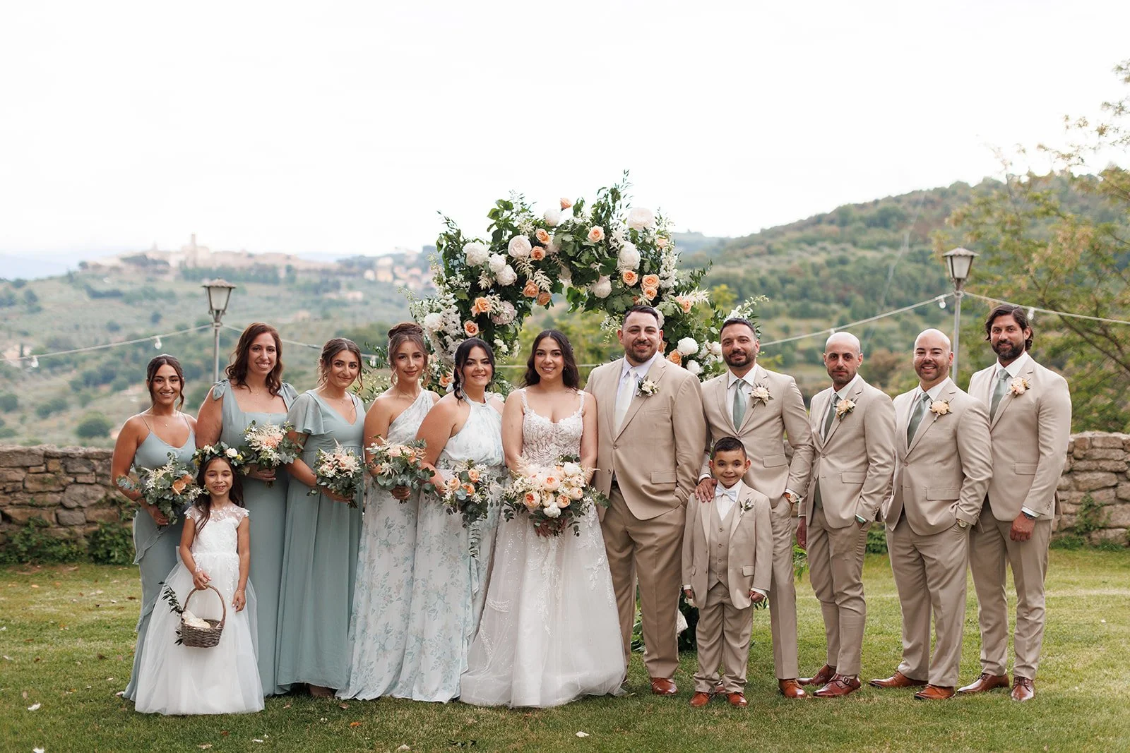 A wedding party outdoors in front of a floral arch with hills in the background. Bridesmaids in light blue dresses, groomsmen and a bride and groom in beige suits. Two young children in white dresses and suits.
