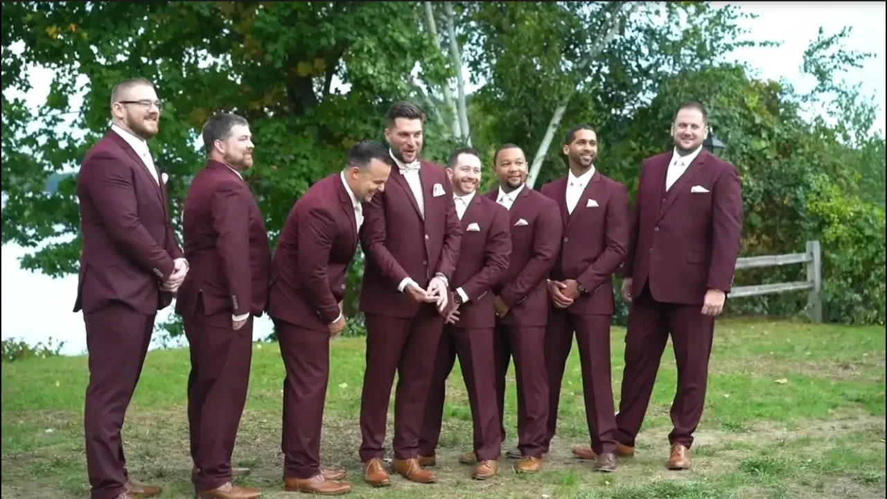 Group of eight men in matching maroon suits standing outdoors on grass, smiling and looking at each other, with trees and a wooden fence in the background.
