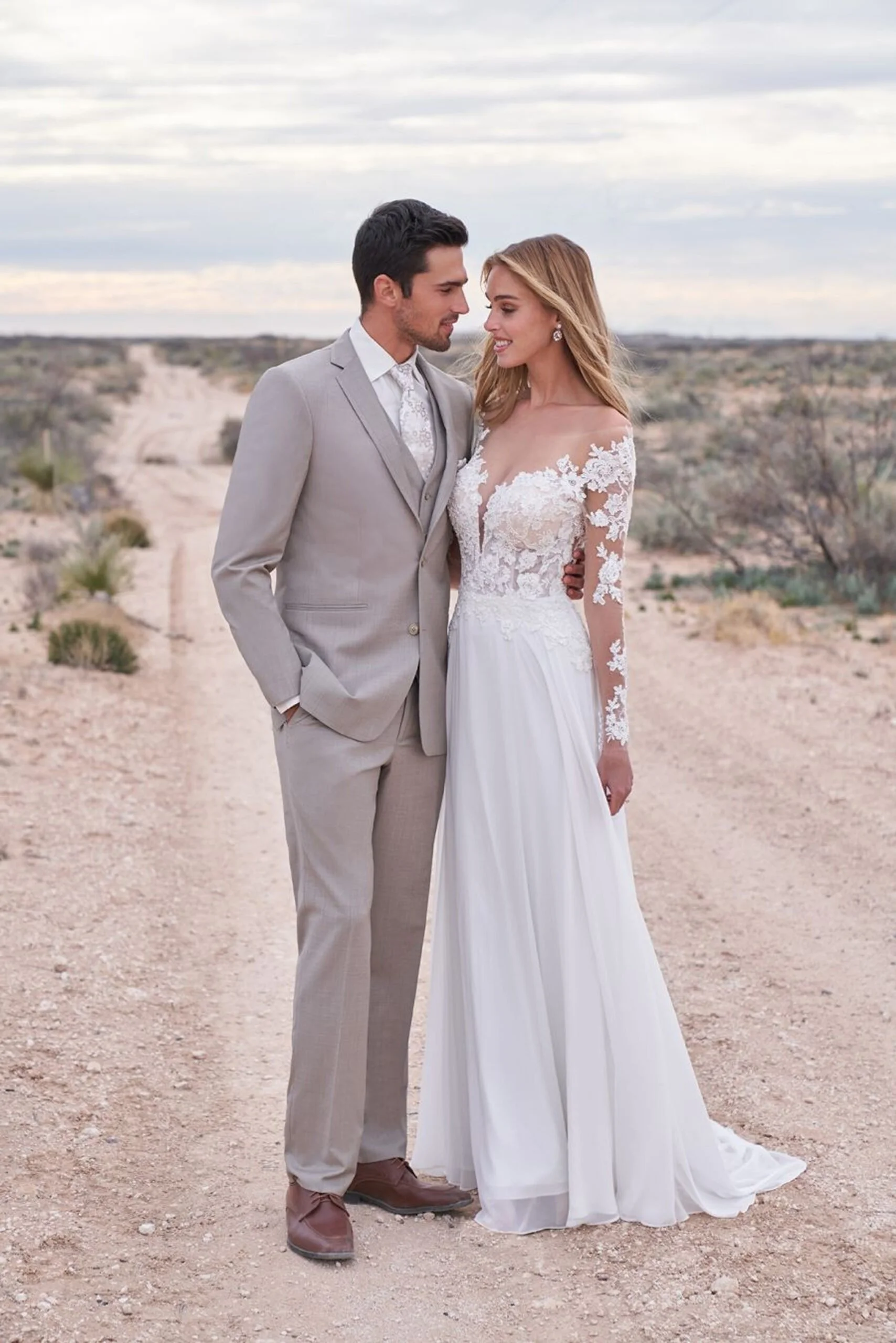 A newlywed couple standing close together on a dirt path in a desert landscape. The groom is wearing a light gray suit with a white shirt and patterned tie, and the bride is in a long white wedding dress with lace details on the shoulders and sleeves. They are smiling at each other with their foreheads almost touching, and the sky is cloudy.