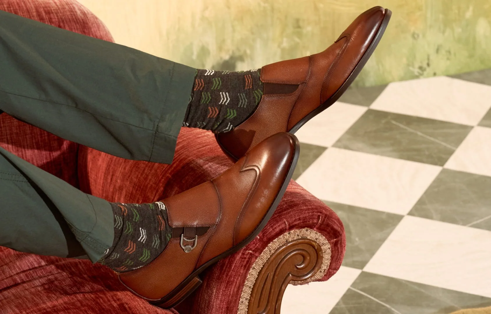 Person wearing brown leather shoes, dark green pants, and colorful patterned socks, sitting on a red upholstered chair with ornate wood trim, on a green and white checkered tiled floor.